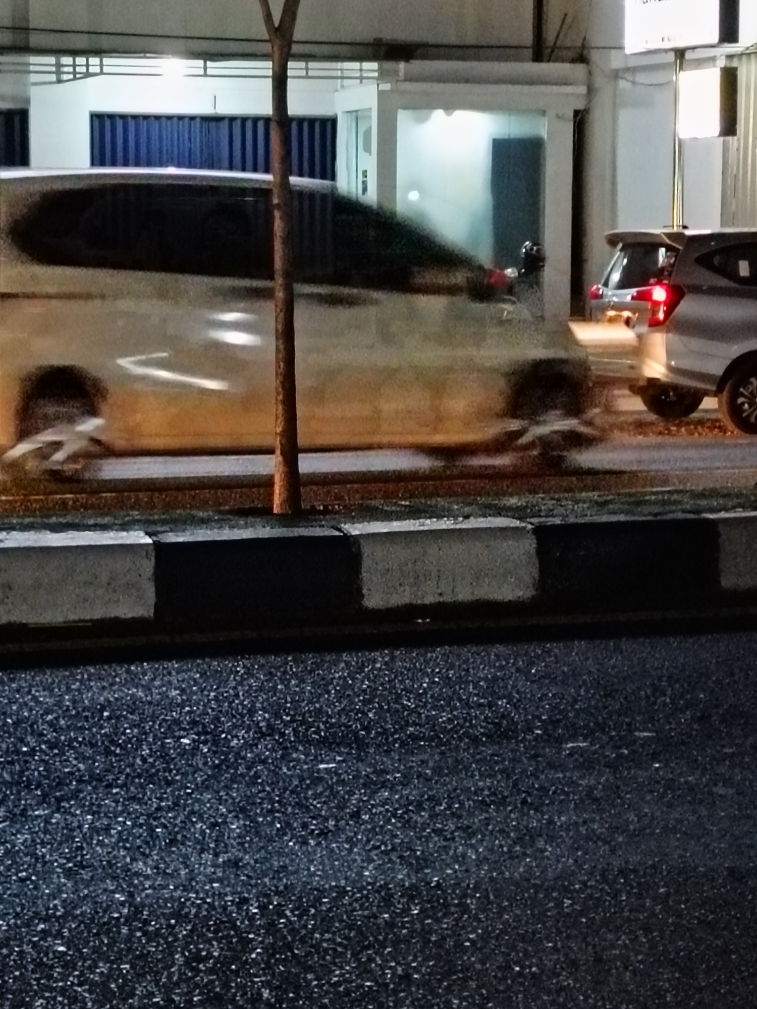 The image captures a nighttime scene on a wet street in Kebondalem Kidul, Indonesia. In the foreground, the dark asphalt road is visible, reflecting faint lights. A concrete curb with black and white markings separates the road from the sidewalk. Beyond the curb, a blurry silver car is in motion, its headlights and taillights creating streaks of light. Partially obstructing the view of the car is a thin tree trunk, also blurred due to motion. In the background, a building with large blue doors and white structures is visible, illuminated by artificial lights. Another car, a silver hatchback, is parked on the right side of the frame, its taillight glowing red. The overall impression is one of a busy street at night, with vehicles in motion and urban infrastructure present.