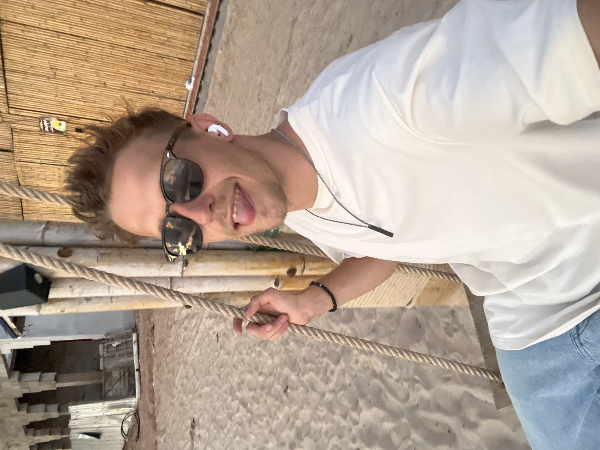 A young Caucasian male with blond, spiked hair and sunglasses is sitting on a swing on a sandy beach. He is wearing a white t-shirt, blue jeans, and white earbuds. He has his tongue sticking out and is holding onto the rope of the swing with his left hand. Behind him, there is a bamboo wall and a building under construction. The image was taken in Choeng Thale, Thailand.