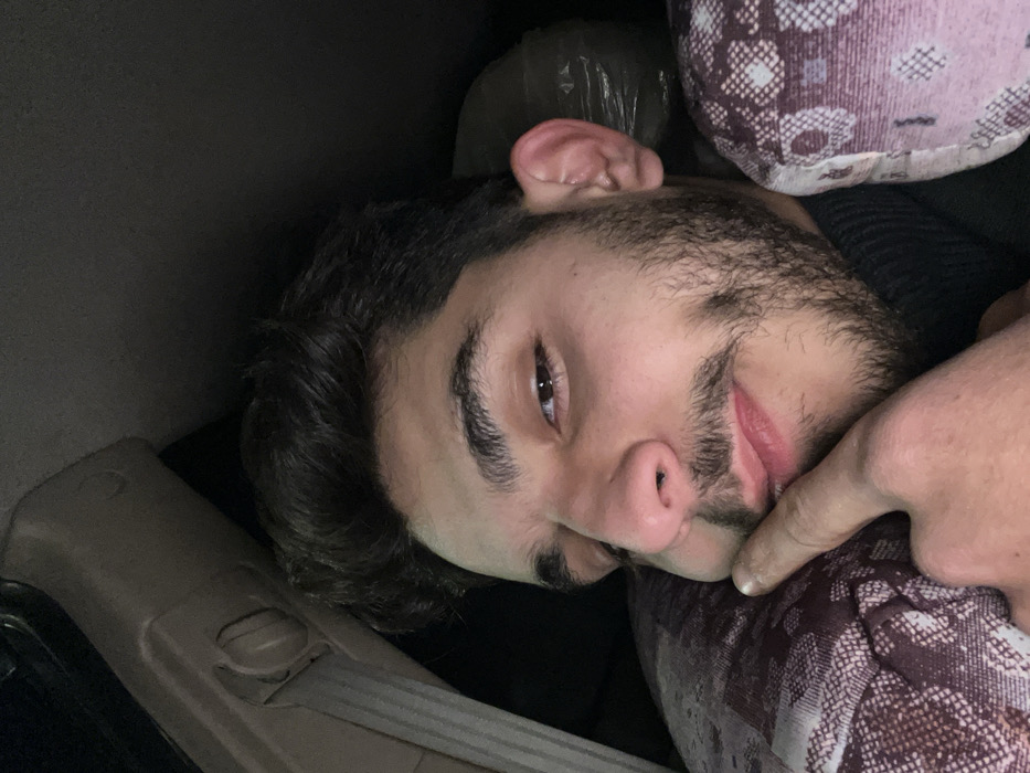 A man with dark hair and a beard reclines against patterned pillows and possibly the interior of a car. He is turned to face the camera with one eye open. His right index finger touches his chin or cheek. A tan interior panel of a vehicle is seen near his head and a dark plastic bag or crumpled material rests behind him. The lighting is low.
