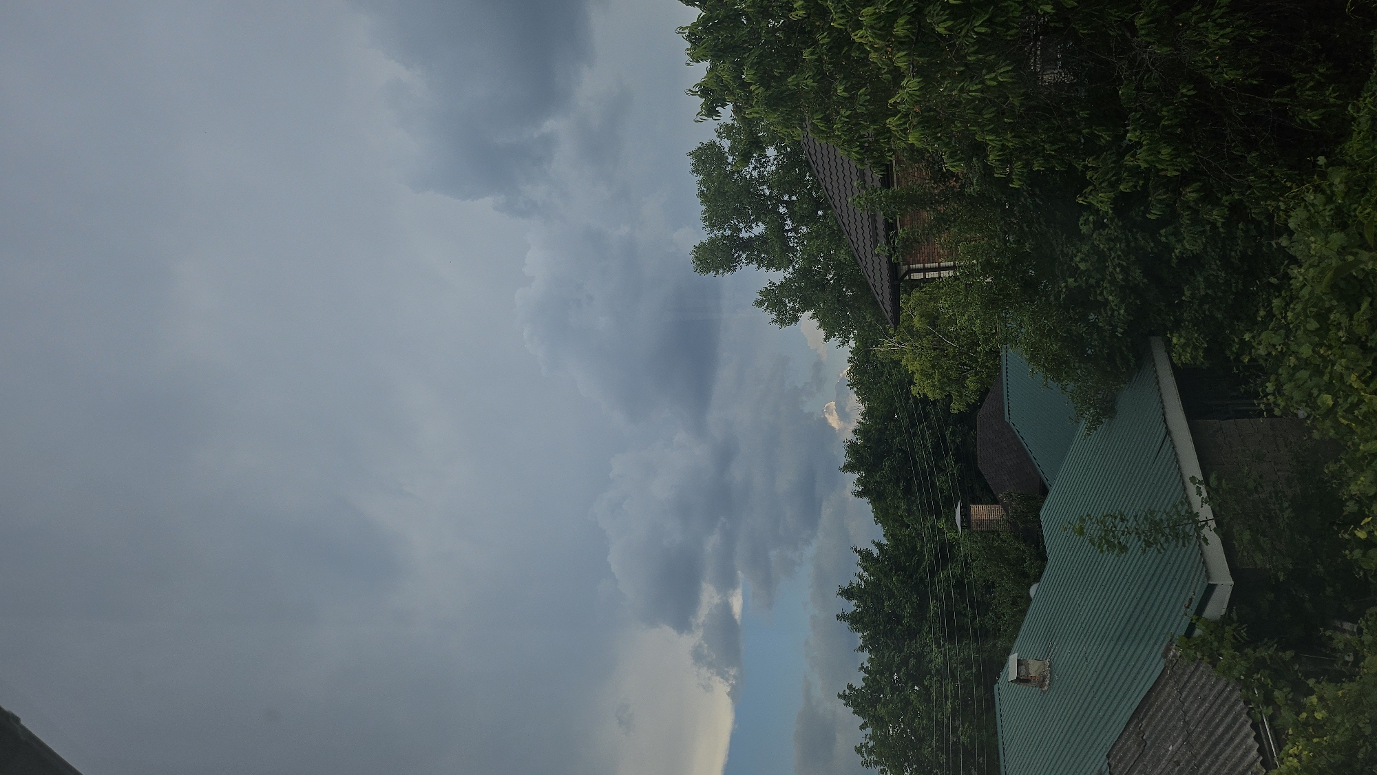This media file displays a cloudy sky above a few houses and lush green trees. The sky is mostly overcast with dark, heavy clouds, suggesting approaching rain or a storm. A sliver of lighter sky and some lighter clouds are visible on the right side of the frame.
The scene is set in a residential area, with the roofs of several buildings partially visible amongst the dense foliage. One house has a dark-tiled roof, while others are topped with green corrugated metal. Chimneys are visible on some of the houses. The trees are full and leafy, indicating a warm season. Power lines are also visible strung between poles.
The lighting suggests it is either early morning or late afternoon, with the overcast sky contributing to a diffused light. No people or specific activities are depicted. The overall mood is somewhat somber due to the dark clouds, but the vibrant green of the trees adds a sense of life to the scene. There is no visible text in the image.