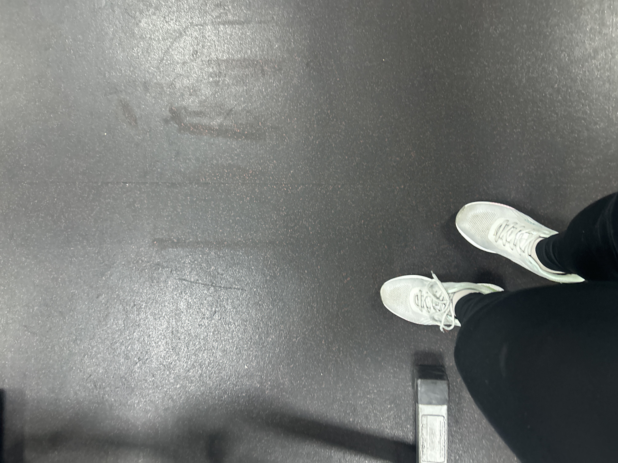 A high-angle, full-body shot taken from a first-person perspective shows a person standing on a dark gray, rubberized floor, commonly found in gyms. The person is wearing black leggings and light gray athletic shoes. To the left of the frame, a portion of a weight-lifting rack is visible, casting a shadow. The floor shows signs of wear, with some scuff marks and lighter speckles. The scene is devoid of any specific identifying markers that would allow for pinpointing its exact location within the city of Omsk, Russia.