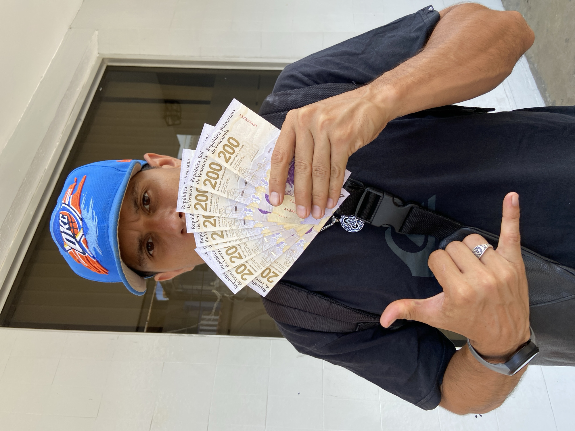 A man stands in an outdoor setting, possibly in San José de Barlovento, Venezuela, holding a fan of Venezuelan Bolivar notes in front of his face. The notes are 200 Bolivar denominations. He wears a blue baseball cap with an orange logo, a black t-shirt, and a black shoulder bag. He is making a hand gesture. He also has a ring on his finger.
