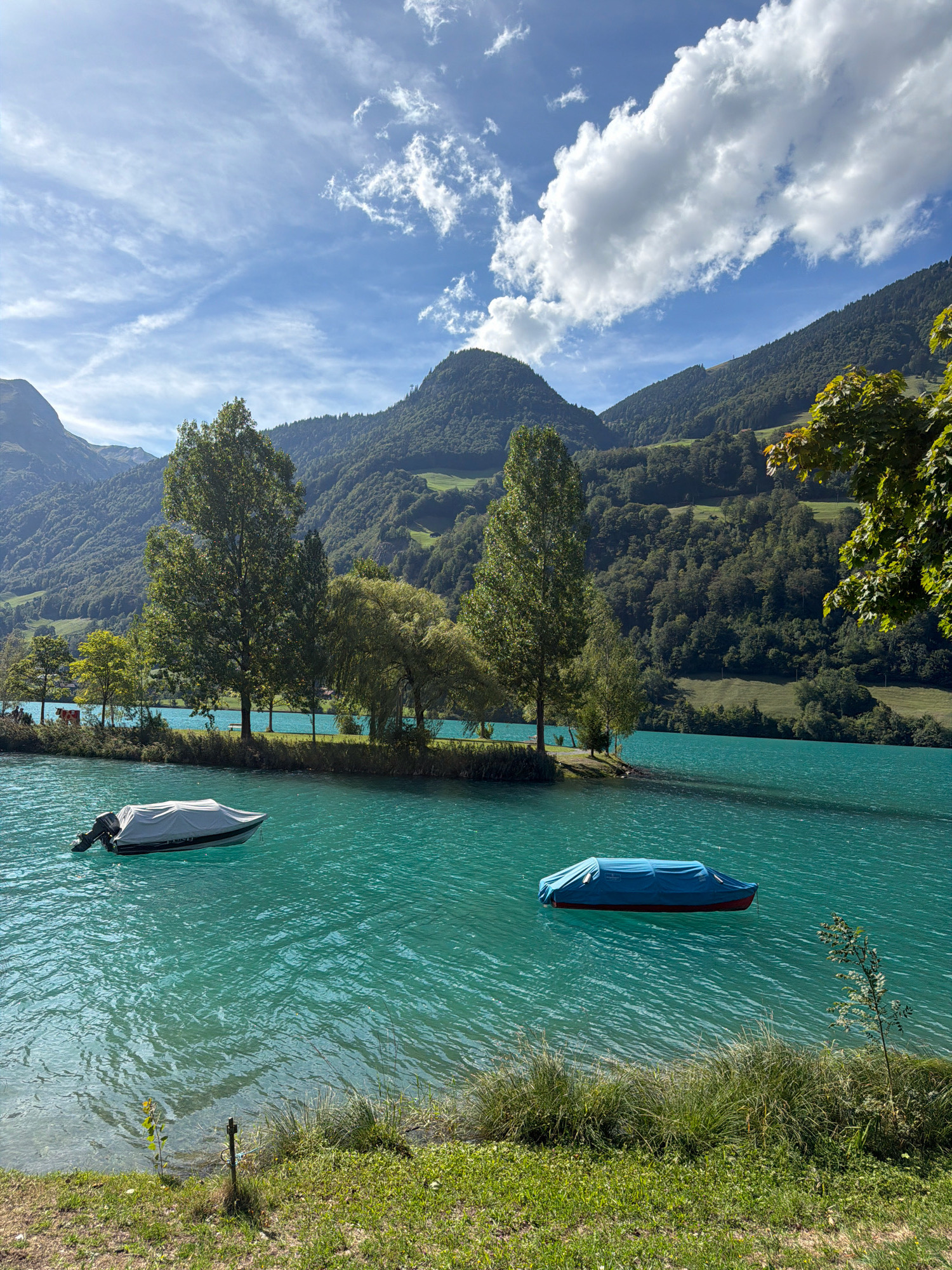 A serene landscape photograph captures a tranquil turquoise lake nestled at the base of lush, tree-covered mountains. The sky above is a vibrant blue, adorned with scattered white clouds. Two boats are visible on the water: one, a motorboat covered with a light gray tarp, drifts on the left side of the lake, while another boat, also covered with a blue tarp, floats on the right. The foreground is a grassy bank, with patches of dry earth and some sparse vegetation. The scene evokes a sense of peace and natural beauty.