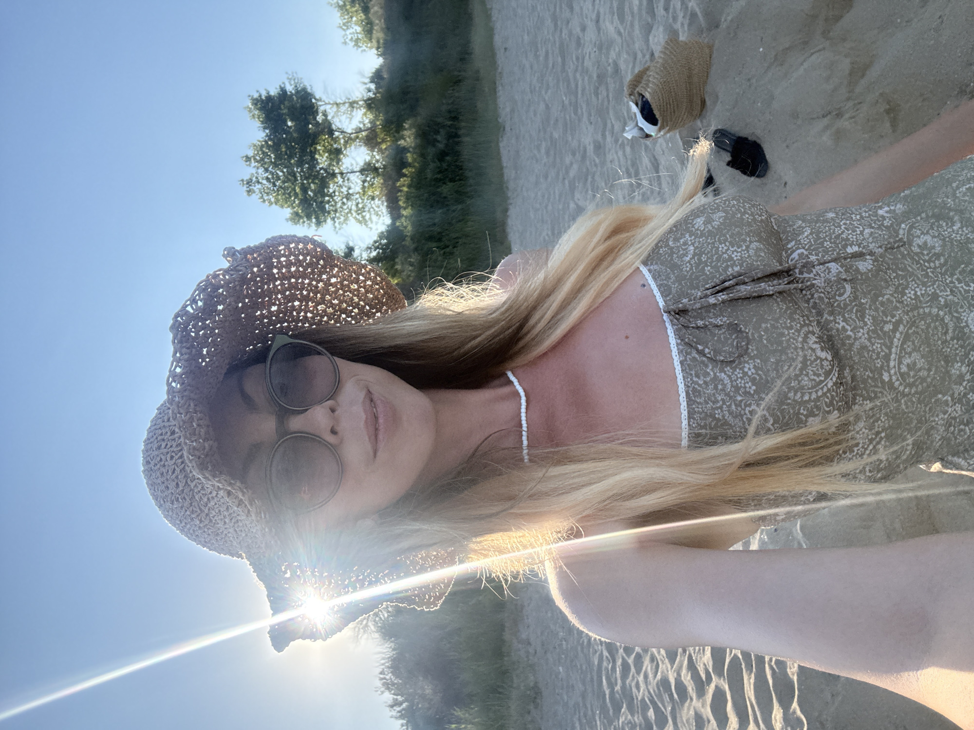 A woman in a straw hat and sunglasses takes a selfie on a sandy beach. She is wearing a light-colored patterned dress and a white beaded necklace. The sun is shining brightly, casting lens flare across the image. In the background, there is a beach, some vegetation, and trees. To her right, a straw bag and a pair of black sandals are visible on the sand. The scene appears to be set in Nesebar, Bulgaria, given the coastal environment and vegetation.