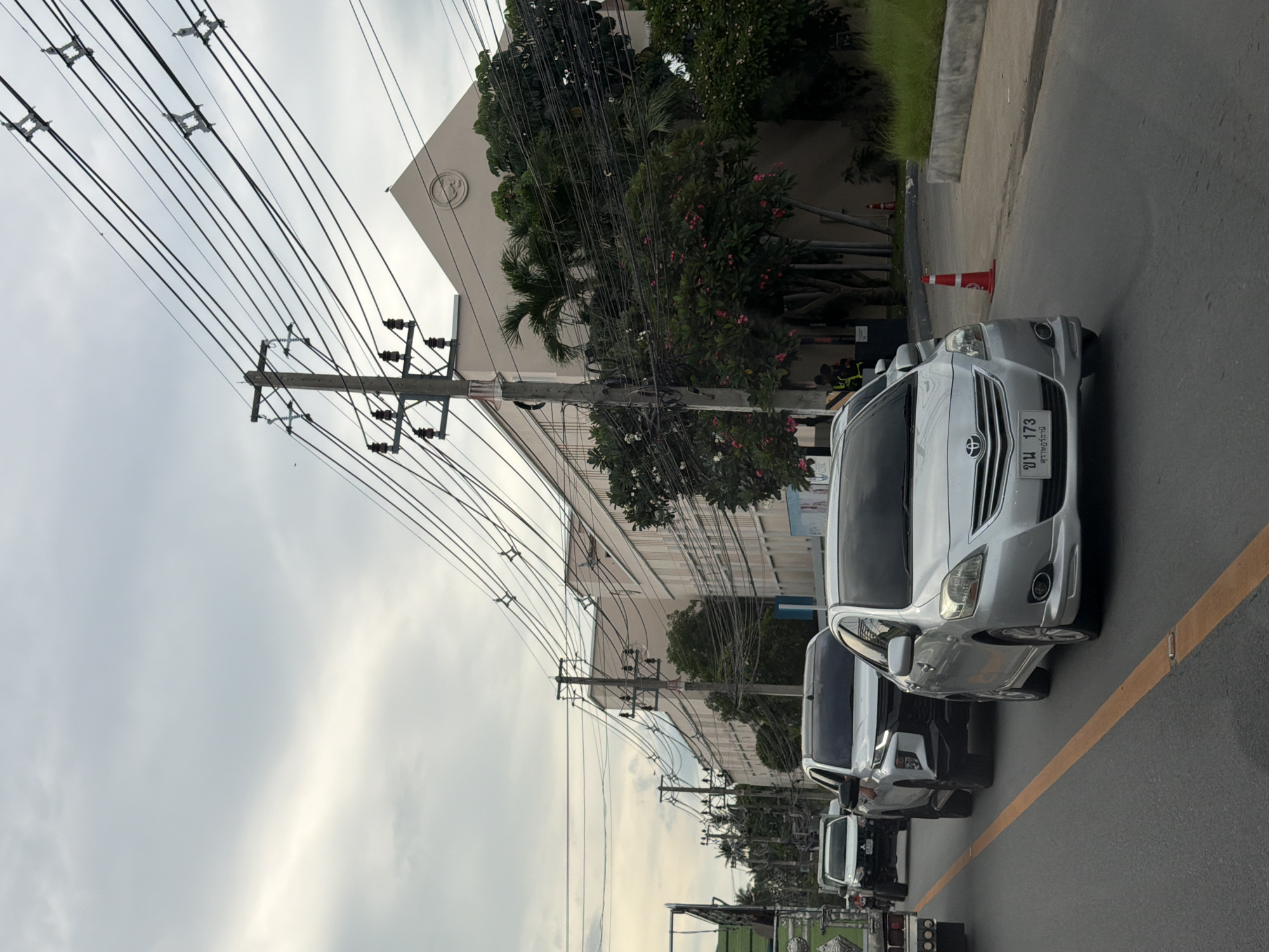 The image captures a street scene in Baan Bo Phut, Thailand, likely during the day, given the ambient light and overcast sky. The foreground features a road with a yellow line marking the lane. Several vehicles are visible, including a silver sedan prominently positioned, displaying a license plate with Thai characters. Behind it, a dark-colored SUV and other vehicles follow, suggesting traffic.
The background is dominated by a long, light-colored building, possibly a commercial or industrial establishment, with trees and palm trees lining its facade. A complex network of overhead power lines stretches across the scene, supported by utility poles, a common sight in many urban and suburban areas. Red and white traffic cones are present on the side of the road, hinting at ongoing work or a temporary traffic diversion. The overall atmosphere appears calm, with no obvious signs of hurried activity. The weather is cloudy, casting a diffused light over the scene.