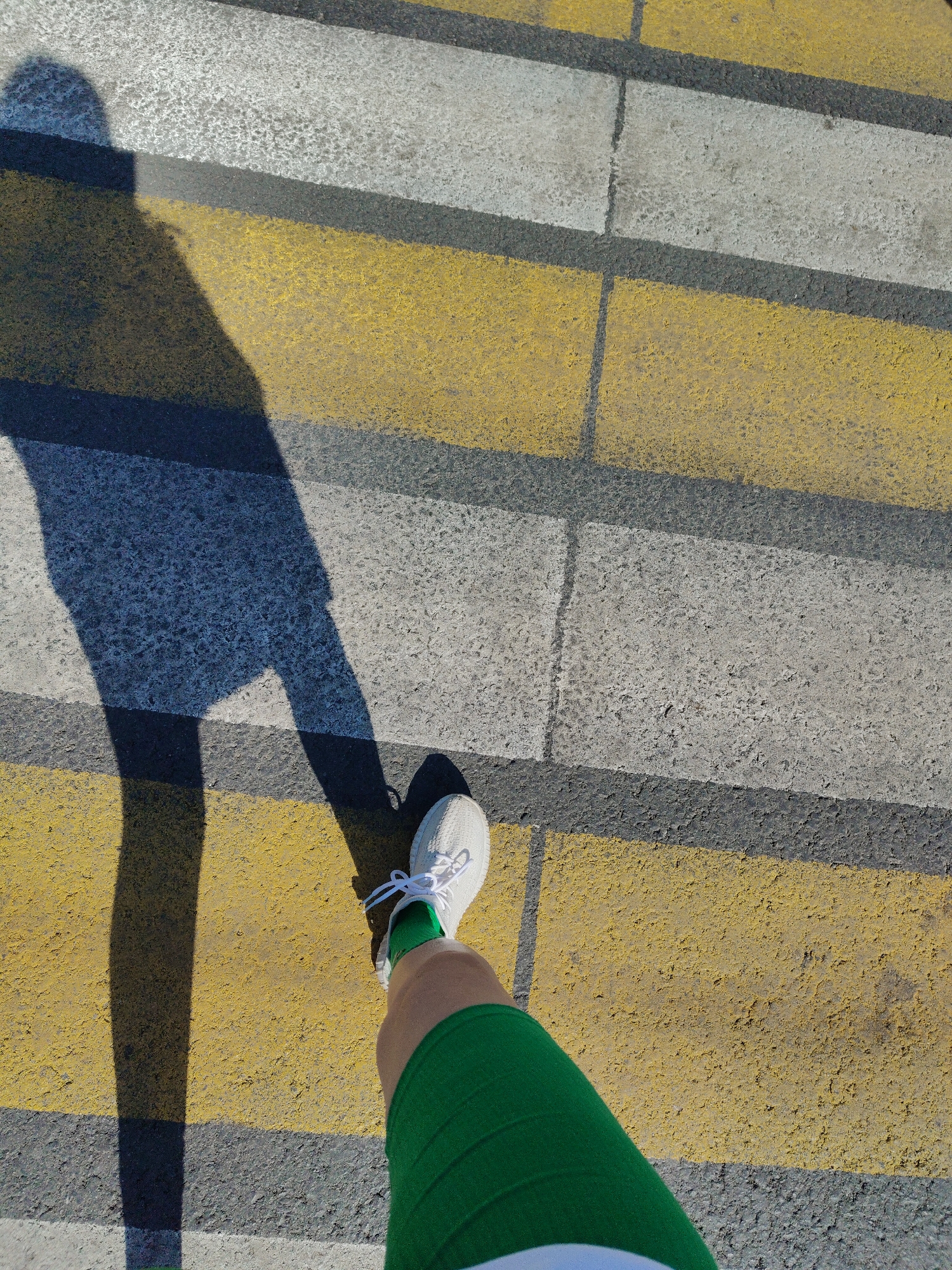 The image shows a person standing on a pedestrian crosswalk in an urban environment. The crosswalk is composed of alternating yellow and white stripes painted on the asphalt surface. The person is wearing a green athletic shorts and white sneakers with visible green socks. The sunlight casts a shadow of the person across the crosswalk. The location appears to be a city street, but the specific city and street are unknown.