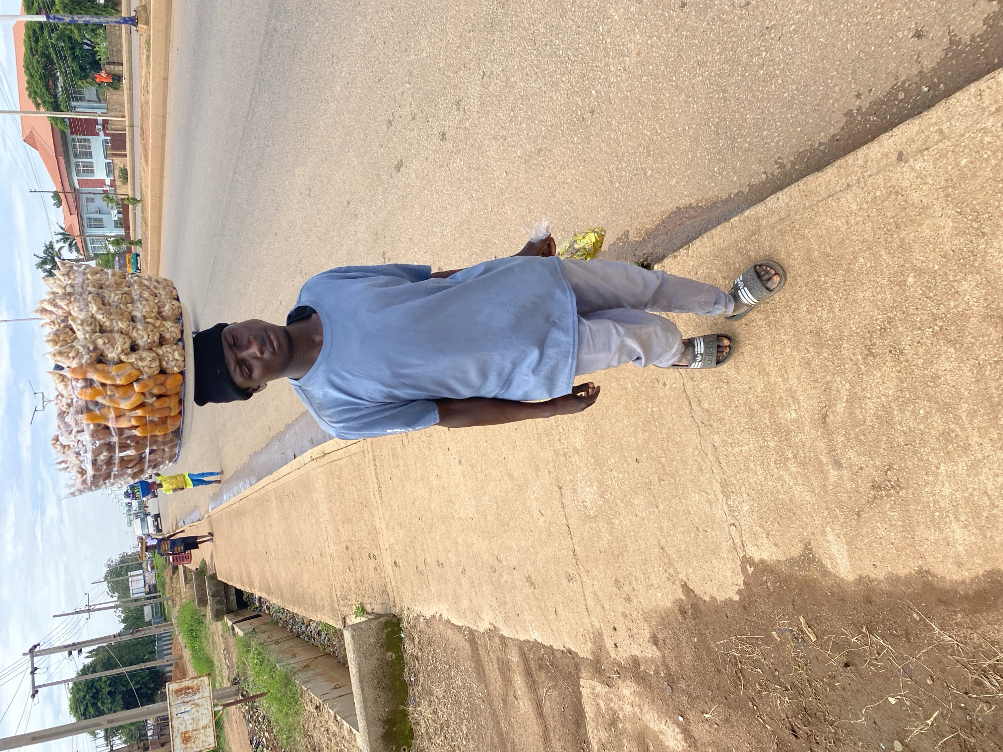 In Bukuru, Nigeria, a young man walks along a dusty concrete sidewalk next to a street. He is wearing a light blue t-shirt, gray pants, and black Adidas slide sandals. He has a black beanie on his head and carries a clear plastic bag with yellow contents in his right hand. Above his head, he balances a large, transparent plastic container filled with what appears to be dried food items, possibly nuts or snacks. The background shows a road with a few vehicles, including a white van and a tricycle, and buildings. A small concrete drainage channel runs along the edge of the sidewalk, filled with dry grass and debris. The sky is partly cloudy.