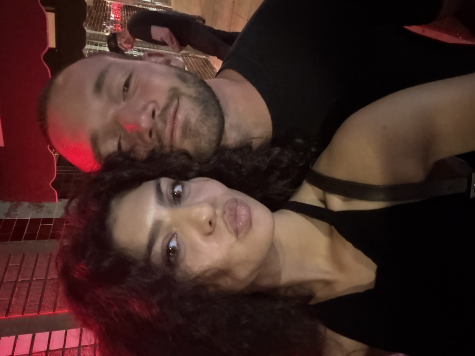 A nighttime scene in West Hollywood, United States of America, depicts two individuals in the foreground posing for a photograph. On the left, an adult female with dark, curly hair and brown eyes is looking directly at the camera with her lips puckered. She is wearing a black halter-neck top, with a dark strap visible over her right shoulder. To her right and slightly behind, an adult male with short dark hair and facial stubble is looking towards the camera with a faint smile. He is wearing a dark crew-neck shirt. A strong red light illuminates the top of the male's head and the left side of the female's hair. In the mid-ground, a dark building facade with visible brickwork or tiling and a red awning is present. In the dimly lit background, a third adult male wearing a dark t-shirt, dark pants, and a dark cap stands further down an urban walkway, facing away from the primary subjects.