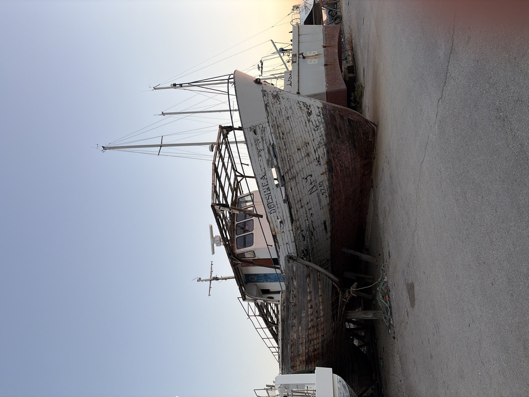 The image depicts an outdoor scene, likely a shipyard or boat storage area, featuring several boats in various states of disrepair. The central focus is a weathered wooden fishing boat with peeling paint, propped up on wooden stands. The boat has a cabin structure with windows, and the name "DESSINEA" is visible on its hull. To the right of the wooden boat, a larger, gray vessel with the number "66" is also visible, resting on similar supports. In the background, the masts of sailboats can be seen against a clear, pale blue sky that transitions to a soft orange hue near the horizon, suggesting either sunrise or sunset. The ground is a paved surface, possibly concrete, with some scattered debris, including ropes and netting, near the base of the wooden boat. The scene appears calm and still, with no people or immediate activity evident. While the prompt specifies a location in Bar, Montenegro, there are no visible landmarks or text within the image that directly confirm this specific geographical placement.