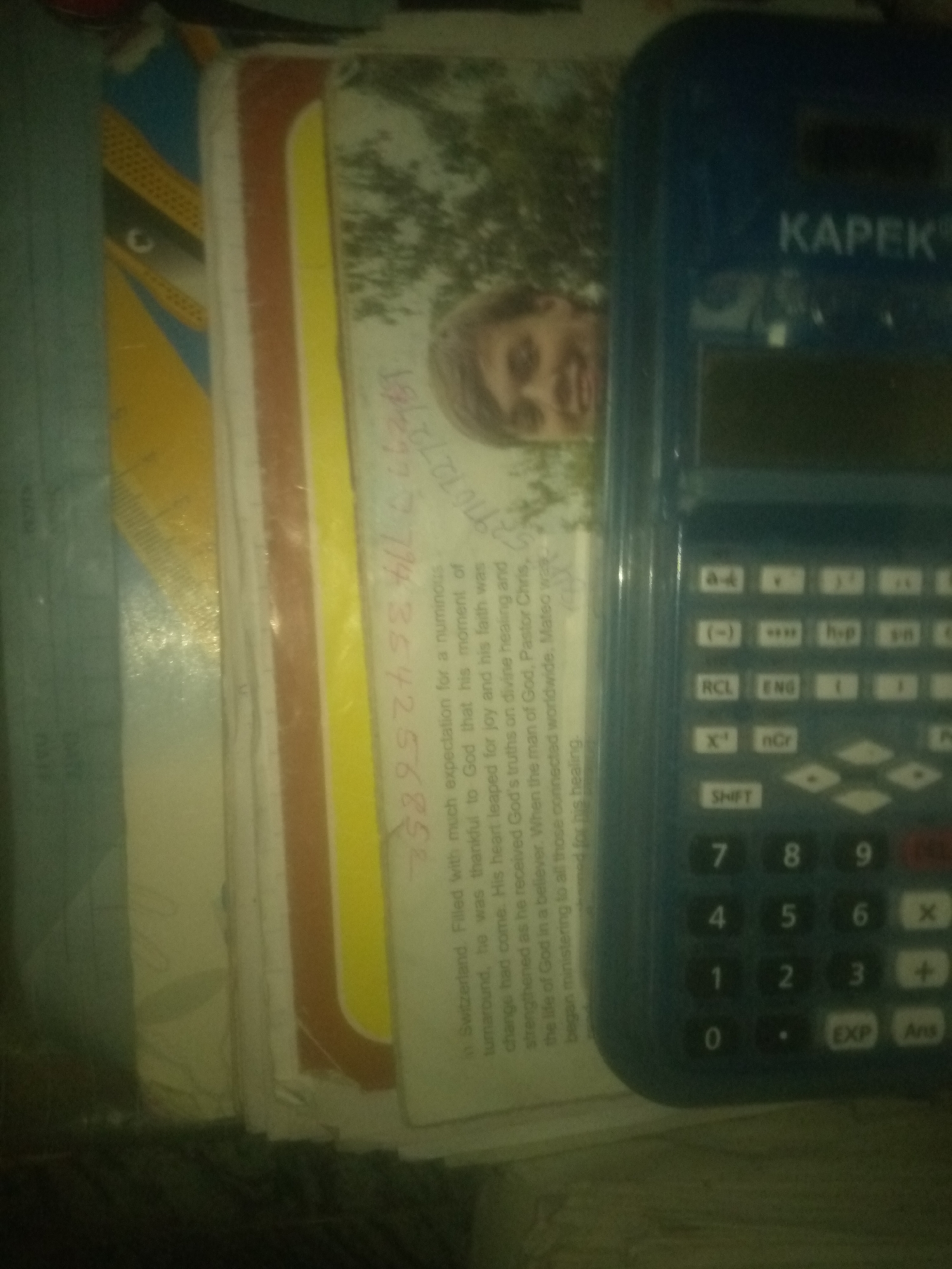 This is a close-up, high-angle, slightly blurred, indoor, still photograph featuring a blue Kapex calculator. The calculator is positioned on the right side of the frame, oriented vertically. Its keypad is visible, showing buttons for numbers 0-9, arithmetic operations, and various functions like "SHIFT", "EXP", and "Ans".
To the left of the calculator, several books or notebooks are stacked. They have different colored spines and covers, with visible text and some illustrations on the pages. One page features a picture of a person's face, looking towards the right. Another page has handwritten text in red ink, with numbers like "27354258". The text on the pages appears to be in English and possibly another language.
The background is dark and out of focus, suggesting an indoor environment. The lighting appears to be dim, with some glare on the calculator's screen. There is no discernible information in the image that can be used to determine the location in the city of Jauro Yinu, Nigeria.