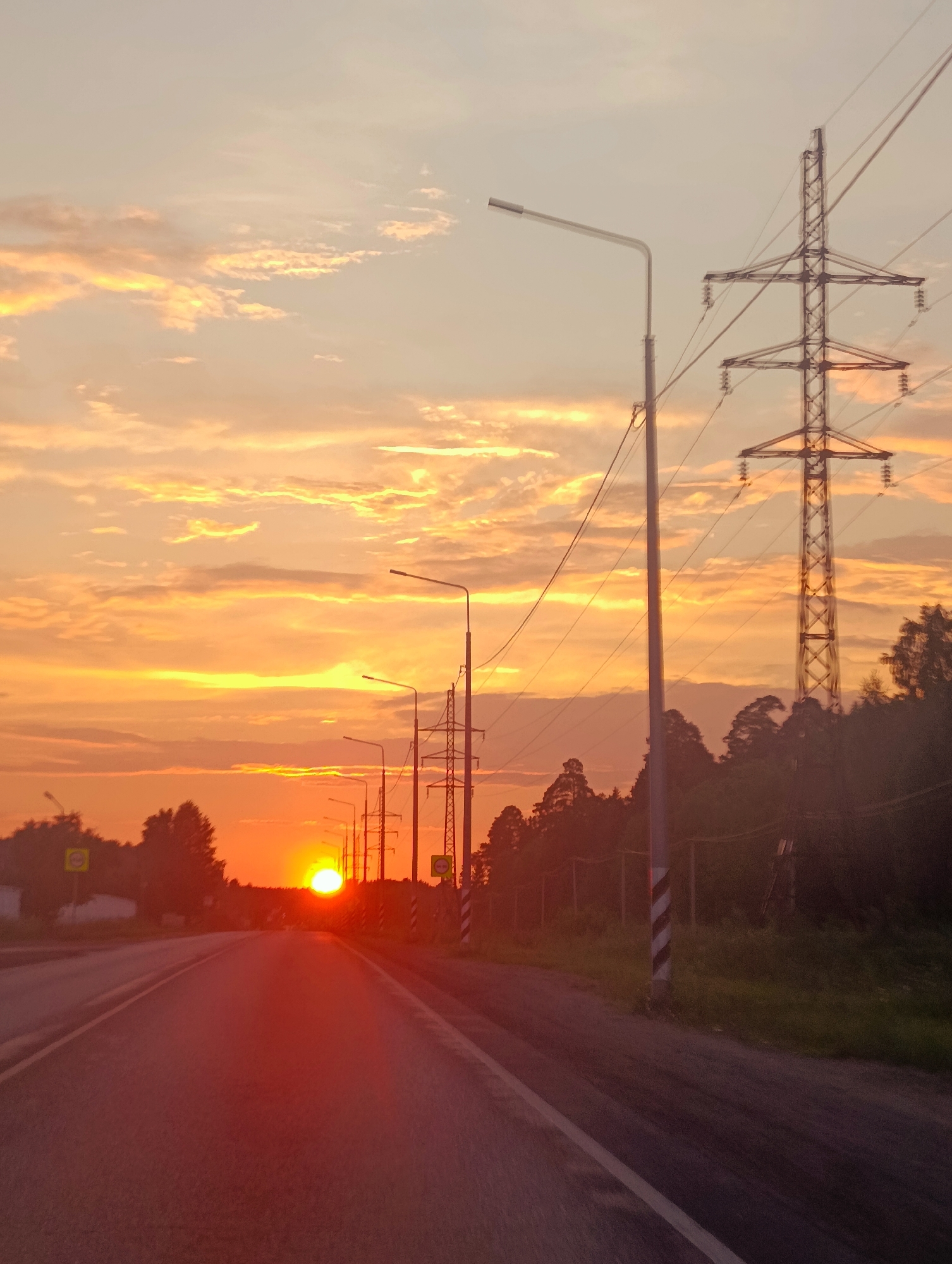 The image depicts a sunset over a road, with a power line tower and streetlights visible. The sun, a bright orange orb, is setting on the horizon, casting a warm glow on the clouds and the surrounding landscape. The road appears to be a highway, with white lines marking the lanes. Trees and a forest can be seen in the background, and a power line tower stands tall against the colorful sky. The scene is set in a rural or suburban area, possibly on the outskirts of Chaikovsky, Russia, given the presence of infrastructure like power lines and streetlights, juxtaposed with the natural beauty of the sunset. There are no people visible in the image.