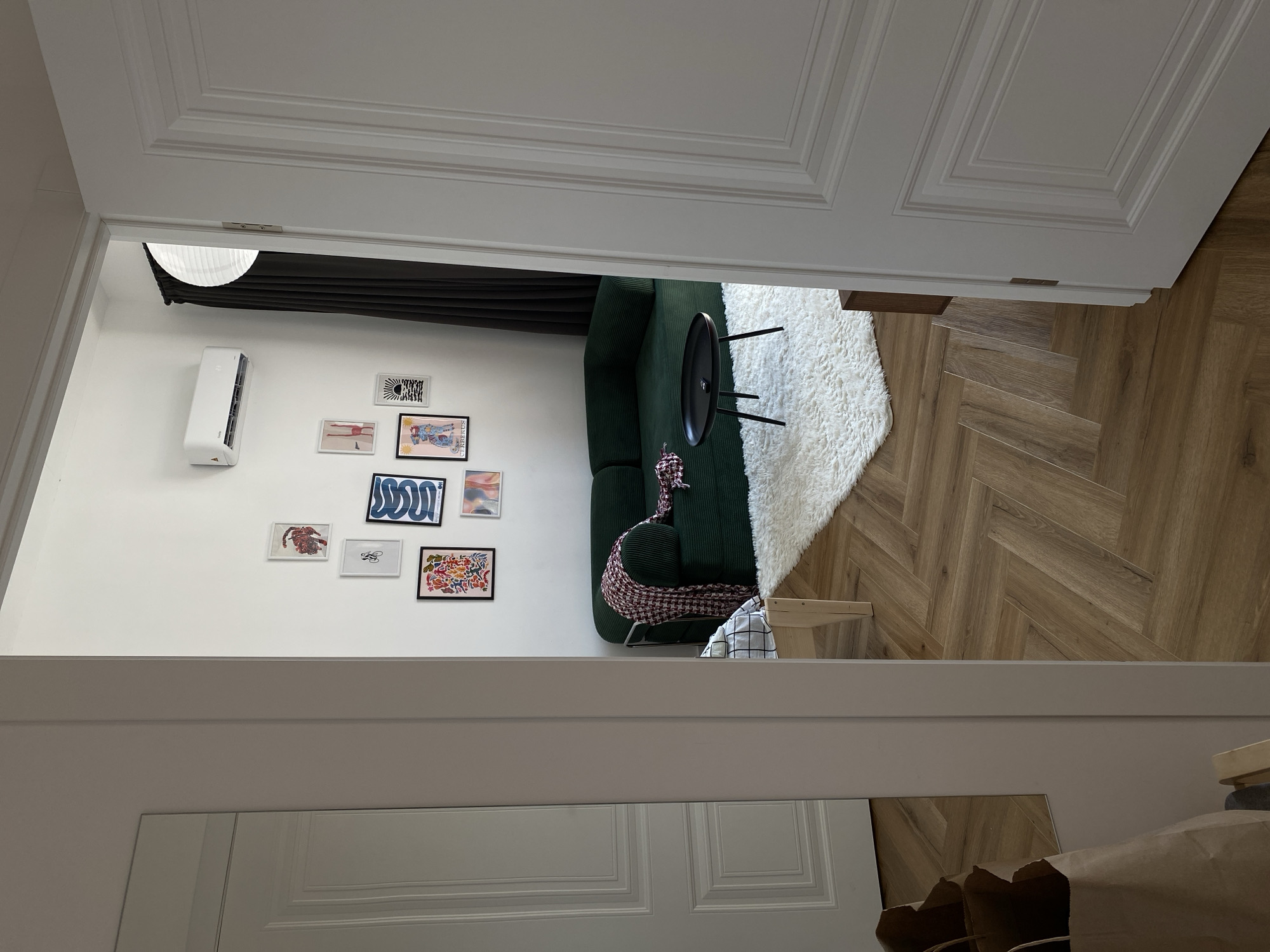 The scene is a view through a partially opened white double door, showing a glimpse into a room. The room has white walls adorned with a collection of small framed pictures. An air conditioning unit is mounted high on the wall. A dark green couch sits in the room. A black circular table stands near the couch, and a white shaggy rug is on the floor. The floor appears to be a herringbone pattern of light brown wood. The bottom of the frame reveals a partial view of what appears to be a large mirror. Bags are visible near the base of the mirror. There are no people visible in the scene. The location is not discernible from the image.