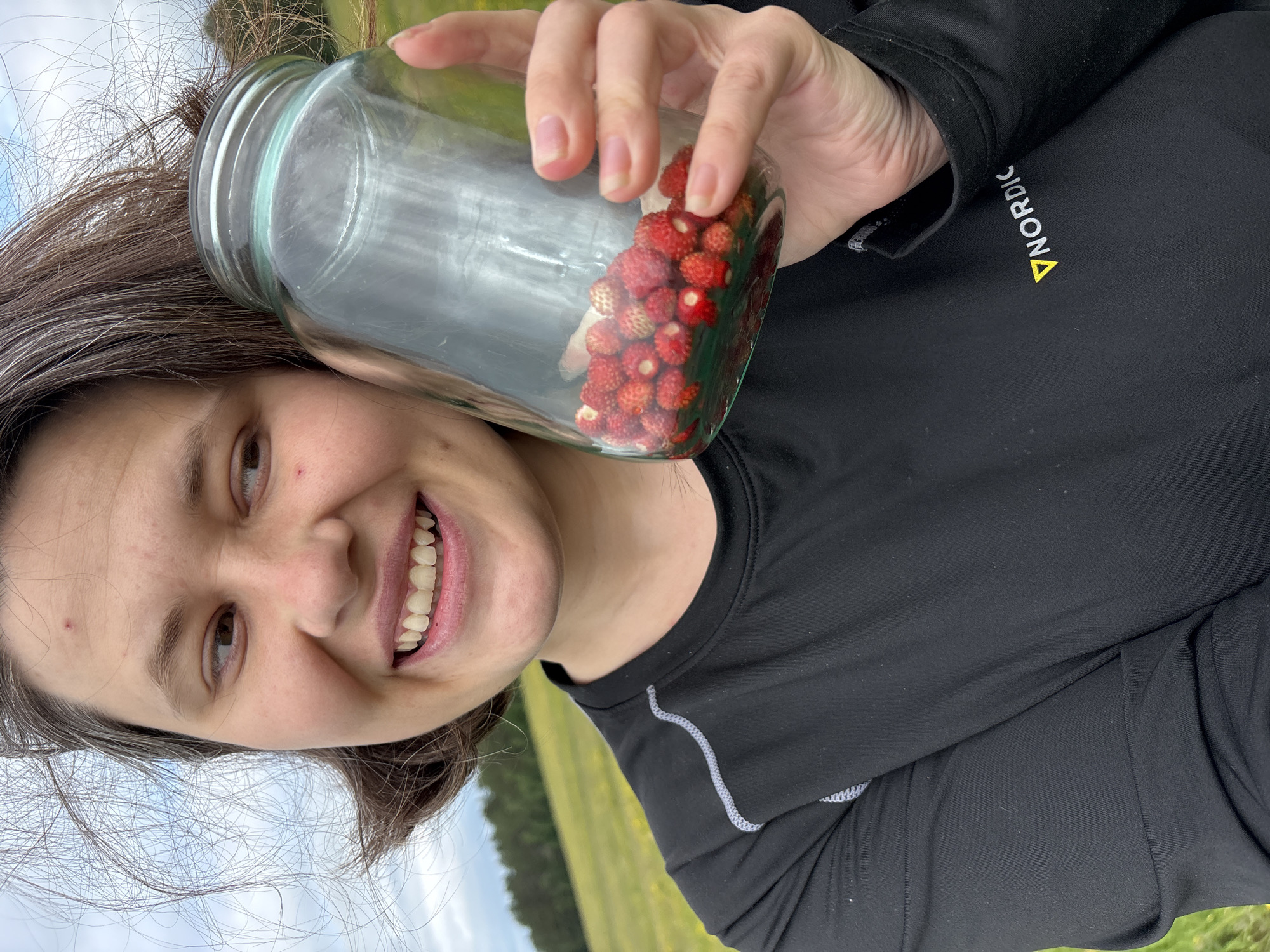 A woman with brown hair, streaked with gray, smiles as she holds up a glass jar filled with small red wild strawberries. She is wearing a black long-sleeved shirt with a white zigzag pattern along the shoulder and a logo that reads "NORDIC" with a yellow triangle. She is outdoors in a grassy field, with trees visible in the background under a cloudy sky. The image appears to be a selfie.
