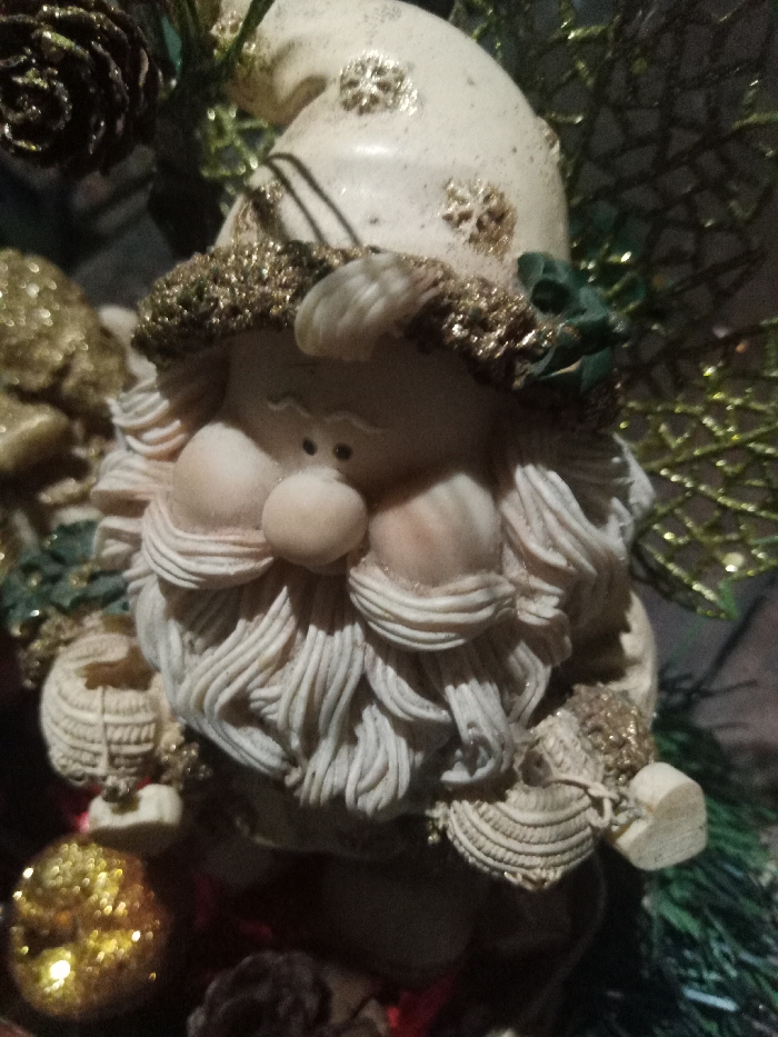 A ceramic Christmas gnome with a textured white beard and a matching hat decorated with gold snowflakes sits amidst holiday decor. The gnome's face is rosy-cheeked with a round nose. Gold glitter trim adorns the hat's brim. Beside the gnome, a shiny gold ornament and a dark pinecone are visible. The background includes green decorative foliage, adding to the festive atmosphere.