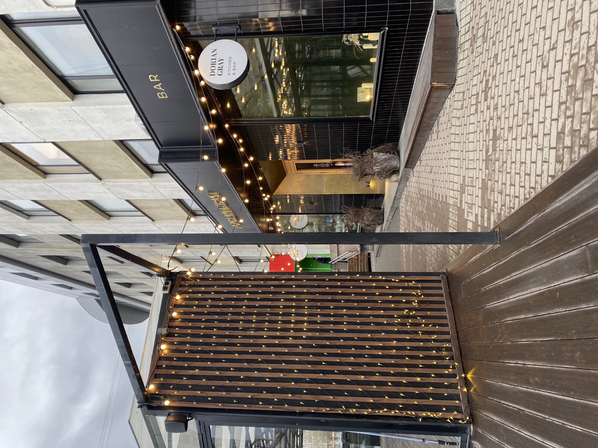 The image captures an outdoor scene in an urban area of Yekaterinburg, Russia, featuring the facade of a building housing a bar and kitchen establishment. The main building on the right has a dark, possibly tiled or paneled, exterior with large windows reflecting string lights. A prominent circular white sign hanging from this building reads "DORIAN GRAY KITCHEN & BAR" in black lettering. Above the main entrance, a black horizontal panel also displays the word "BAR" in gold capital letters.
In front of the establishment, there is an outdoor patio or seating area constructed from dark wooden slats and a black metal frame. Numerous warm-toned string lights are illuminated, draped across the building's facade and woven through the wooden slats of the outdoor structure, creating an inviting ambiance. The ground is paved with light-colored bricks, visible extending to the right. To the left, another building with a lighter, possibly concrete or stucco, facade and dark-framed windows is partially visible. The sky is overcast, and no people are present, indicating a quiet moment. Small potted plants with dried branches are situated near the entrance of the bar.