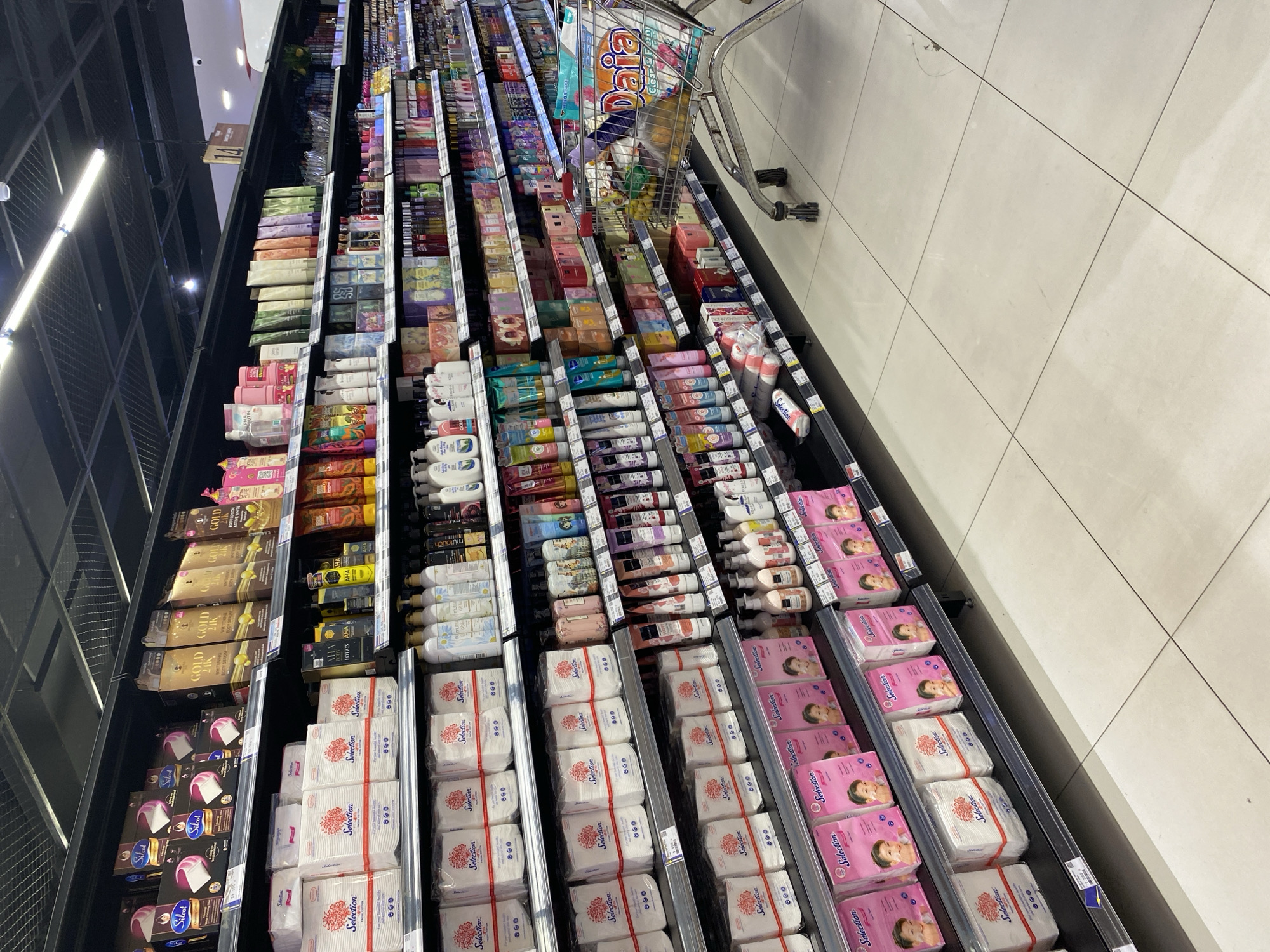 A wide-angle shot captures an aisle in a brightly lit supermarket in Banda Aceh, Indonesia. The shelves are stocked with a variety of personal care products, including facial tissues, body lotions, shampoos, and soaps. The products are neatly arranged, with brands like "Selection," "Gold 24K," and "AHA" visible. The aisle is clean and well-maintained, with tiled flooring. A shopping cart is positioned in the foreground, partially filled with grocery items. The overall impression is one of a well-organized and well-stocked retail environment.