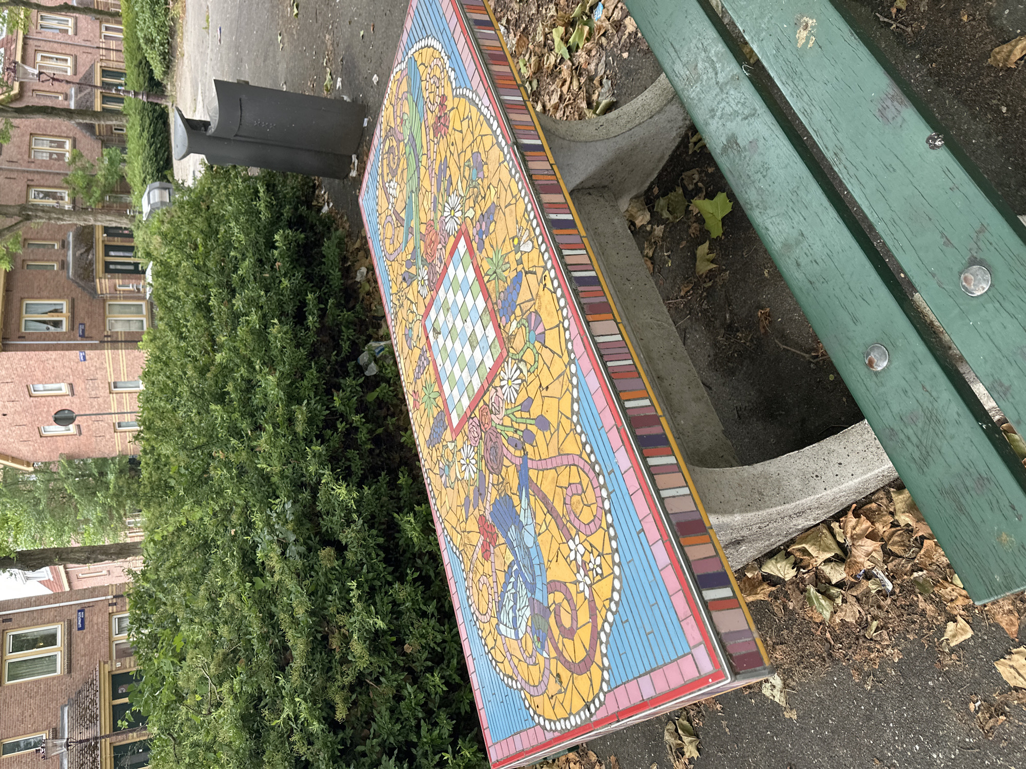 The image depicts a mosaic-tiled bench in an outdoor urban setting. The bench features a colorful mosaic design with intricate patterns, including what appears to be a chessboard in the center, surrounded by floral and bird motifs. The bench itself is made of concrete with decorative mosaic tiles along its edges. In the foreground, a green wooden bench is partially visible. The background shows residential buildings with brick facades, trees, and a dense green hedge. A dark-colored trash can is also present. The ground is paved with asphalt and scattered with dry leaves, indicating an outdoor public space, possibly a park or courtyard. The buildings appear to be in the city of Amsterdam, Netherlands, suggested by the architectural style. There are no people visible in the scene.