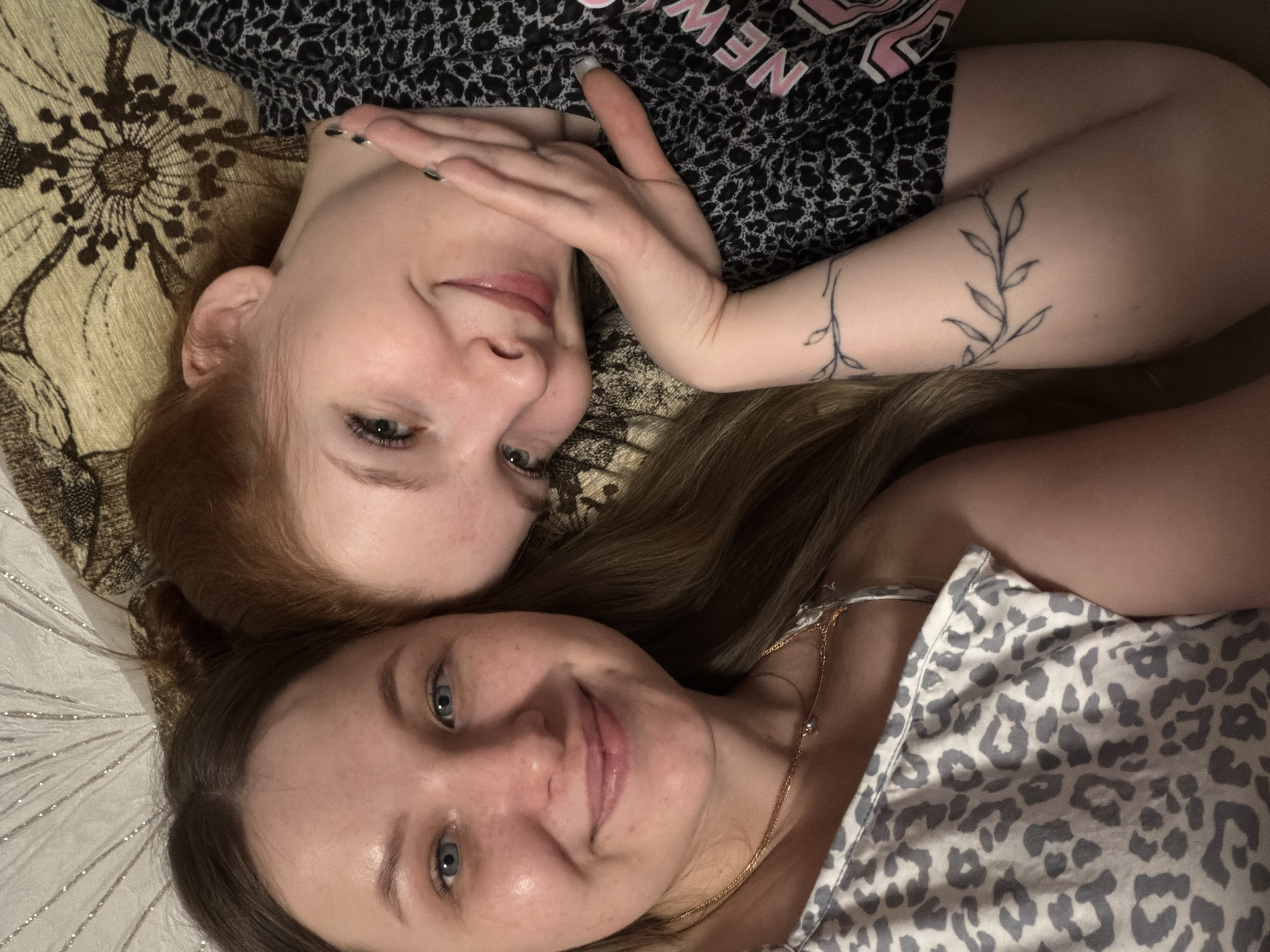 Two women are lying down, facing the camera. The woman on the left has red hair and fair skin. She is wearing a black and white patterned top with pink lettering. Her right hand is near her face and her left arm is visible, with a tattoo of a vine with leaves. The woman on the right has long brown hair and fair skin. She is wearing a light gray and white patterned top and a gold necklace. They are lying on patterned fabric that appears to be a bed or couch. The exact location in the city of Chaikovsky, Russia cannot be determined based on the provided image.