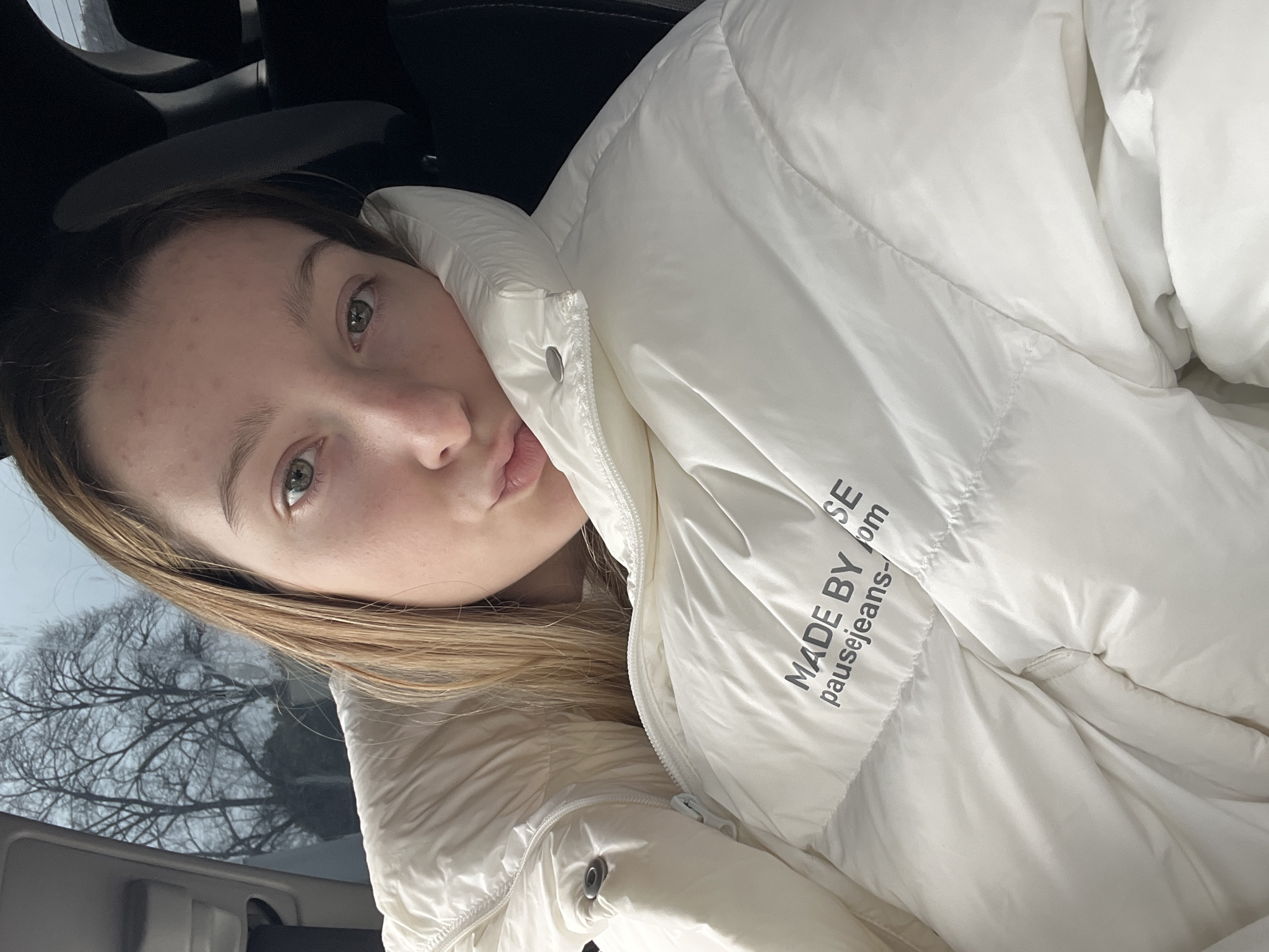 The image shows a woman wearing a white puffer jacket with the text "MADE BY pausejeans-com" printed on it. She is inside what appears to be a car, based on the visible car interior elements. Through the window, bare trees can be seen. The image does not provide enough information to determine the specific location in Varna, Bulgaria.