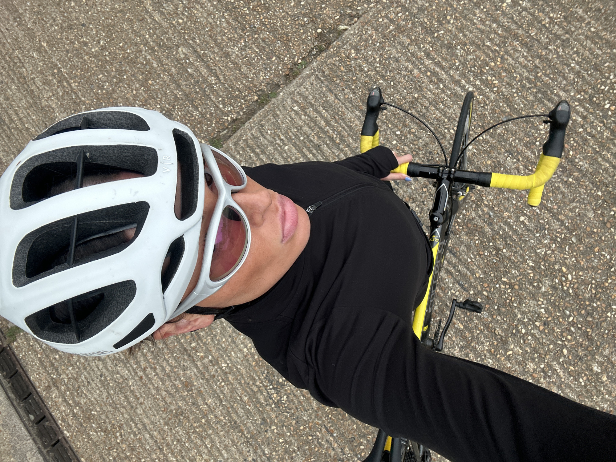 A cyclist wearing a white helmet and pink tinted sunglasses is positioned on a road or path, likely in the city of Tonbridge and Malling, United Kingdom. The person is wearing a long-sleeved black top and holding the handlebars of a bicycle, which is yellow and black. The surface beneath them appears to be a concrete or paved pathway. The view is taken from above, looking down at the subject.