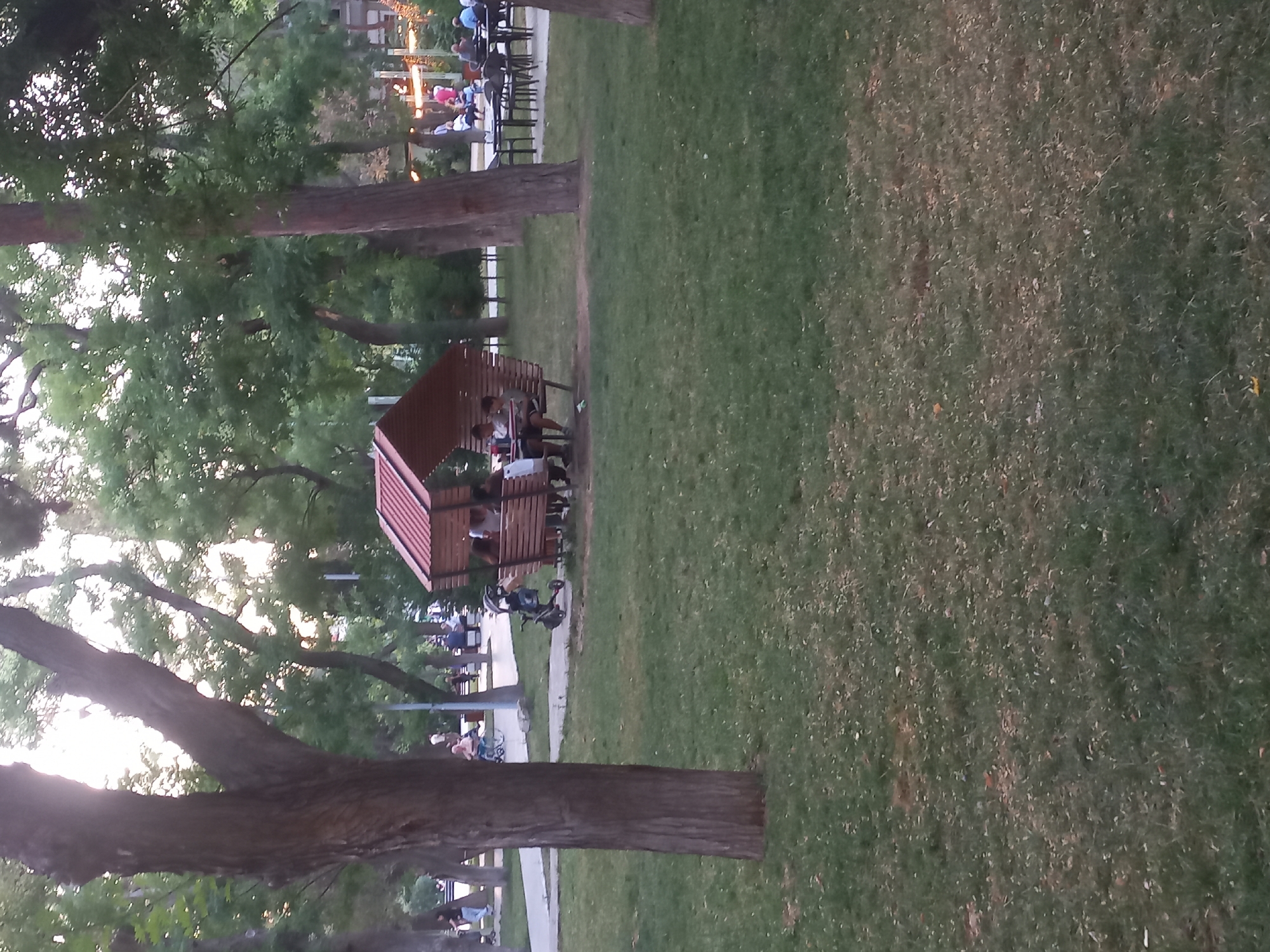 The image depicts a park scene with a small wooden gazebo providing shade for several individuals. The gazebo has a reddish-brown, slanted roof and open sides, with wooden benches inside where people are seated. One person is visible in the foreground of the gazebo, while others are further back. A stroller is parked near the gazebo. In the background, there are more trees, a paved walkway, and other park amenities, including tables and chairs set up outdoors. Several people are scattered throughout the park, some walking and others sitting. The overall environment suggests a public park with green grass and mature trees. Based on the architectural style of the gazebo and the overall atmosphere, this scene is likely located in a city park within Baku, Azerbaijan.