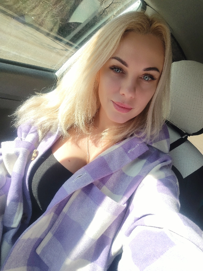 A blonde woman with blue eyes is depicted in a selfie taken inside a vehicle. She is looking directly at the camera with a subtle smile. She wears a black top under an open purple and white checkered flannel shirt. A slender gold-colored necklace is visible. Bright sunlight illuminates her from the left, highlighting her hair and face. A car window and roof lining are visible in the upper left background, while a light-colored car seat headrest is partially visible to the right. The location is Орлиновский округ, Ukraine.