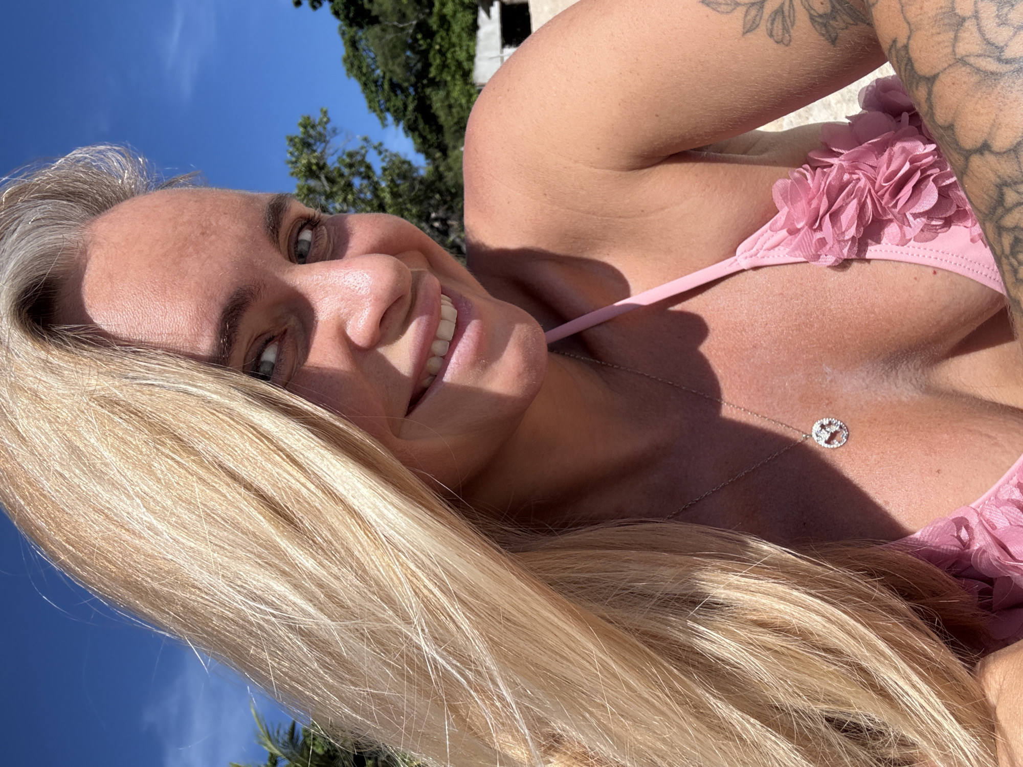 In Baan Chaweng Noi, Thailand, a close-up, waist-up selfie is captured. The subject, a woman with blonde hair, smiles into the camera. She is wearing a pink bikini top adorned with floral embellishments and a silver necklace with a heart pendant. The background shows a clear blue sky with scattered clouds, and the top of lush green trees. To the far right of the image, part of a building is visible. The woman has tattoos on her right arm and shoulder. The sunlight suggests it is daytime.