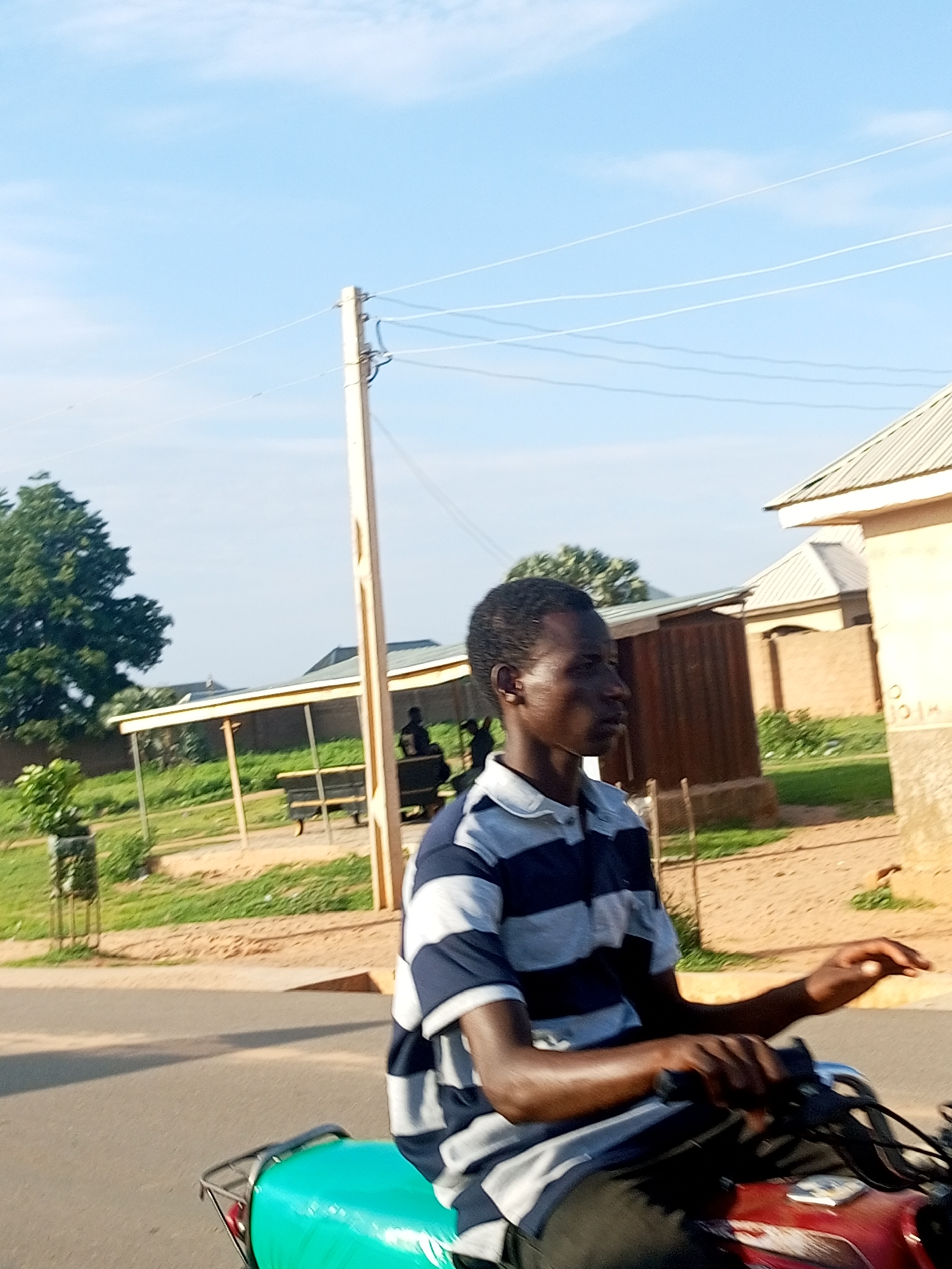 The image shows a young man riding a motorcycle on a dirt road in a village setting. The man is wearing a blue and white striped shirt and black pants. He is looking to the left, with his hands on the handlebars. The motorcycle is red and has a green seat. In the background, there are several buildings, including a house with a corrugated metal roof and a building with a thatched roof. There are also trees and a utility pole with power lines. The sky is clear and blue. The location is identified as Tempure, Nigeria.