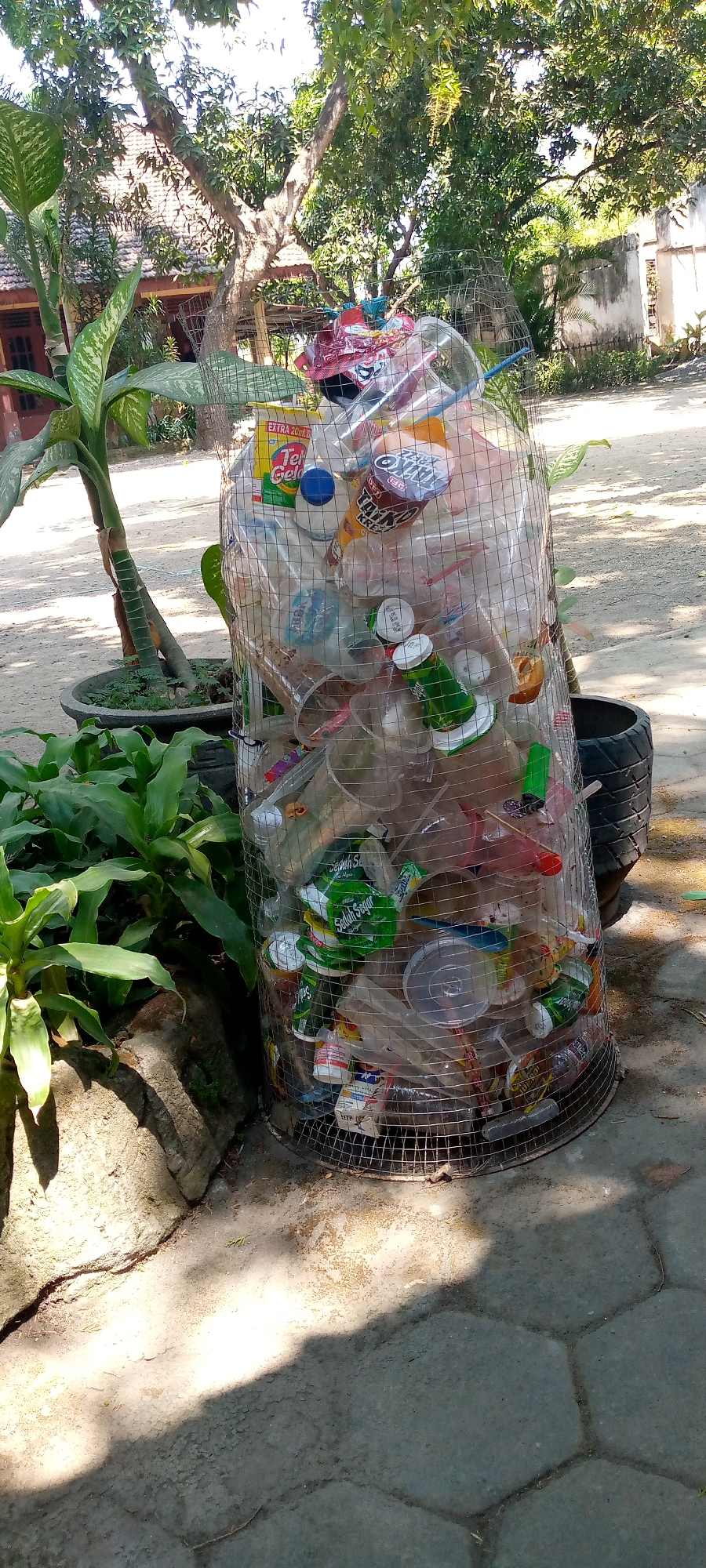 The image shows a cylindrical wire mesh trash bin filled with various plastic waste, including bottles, cups, and wrappers. The bin is situated outdoors on a paved surface, possibly in the city of Pacing, Indonesia. Lush green foliage is visible to the left of the bin, and a large tree with dense leaves provides shade overhead. In the background, part of a building with a tiled roof and a wall can be seen. The lighting suggests it is daytime, with sunlight casting shadows on the ground. No people are present in the image.