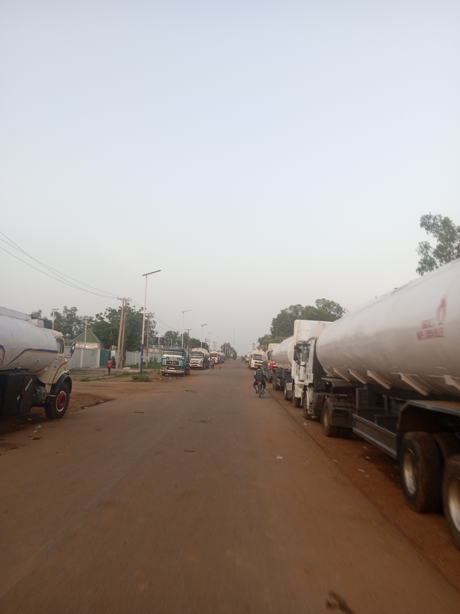 The image captures a street scene in Television, Nigeria, characterized by a long queue of tanker trucks. The trucks, primarily white, are parked bumper-to-bumper along the right side of a reddish-brown dirt road. Some of the trucks have their reflective markers or company logos visible. In the foreground on the left, a portion of a cream-colored tanker truck is visible, showing its wheel and part of its chassis. The road extends into the distance, where more tanker trucks and various other vehicles, including what appears to be a motorbike with a rider, are present.
On the left side of the road, there are buildings with green roofs, possibly residential or commercial structures, bordered by a gray wall. Several utility poles with wires run along the street, and some modern streetlights are also visible. Trees and greenery are scattered throughout the scene, suggesting an urban or semi-urban environment. The sky is overcast with a light, hazy gray color. A few individuals are visible walking near the buildings on the left, and one person is seen riding a motorbike on the left side of the road. The overall impression is one of a significant traffic congestion or a waiting line of fuel tankers.