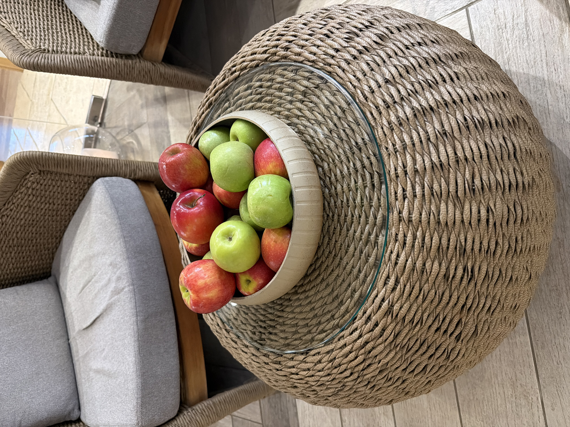 A round wicker coffee table with a glass top sits in front of two chairs. A bowl filled with apples, a mix of red and green, is placed on the coffee table. The chairs have gray cushions. The background shows a hallway with tiled flooring and a glass door. The location is Galil Elyon Regional Council, Israel.