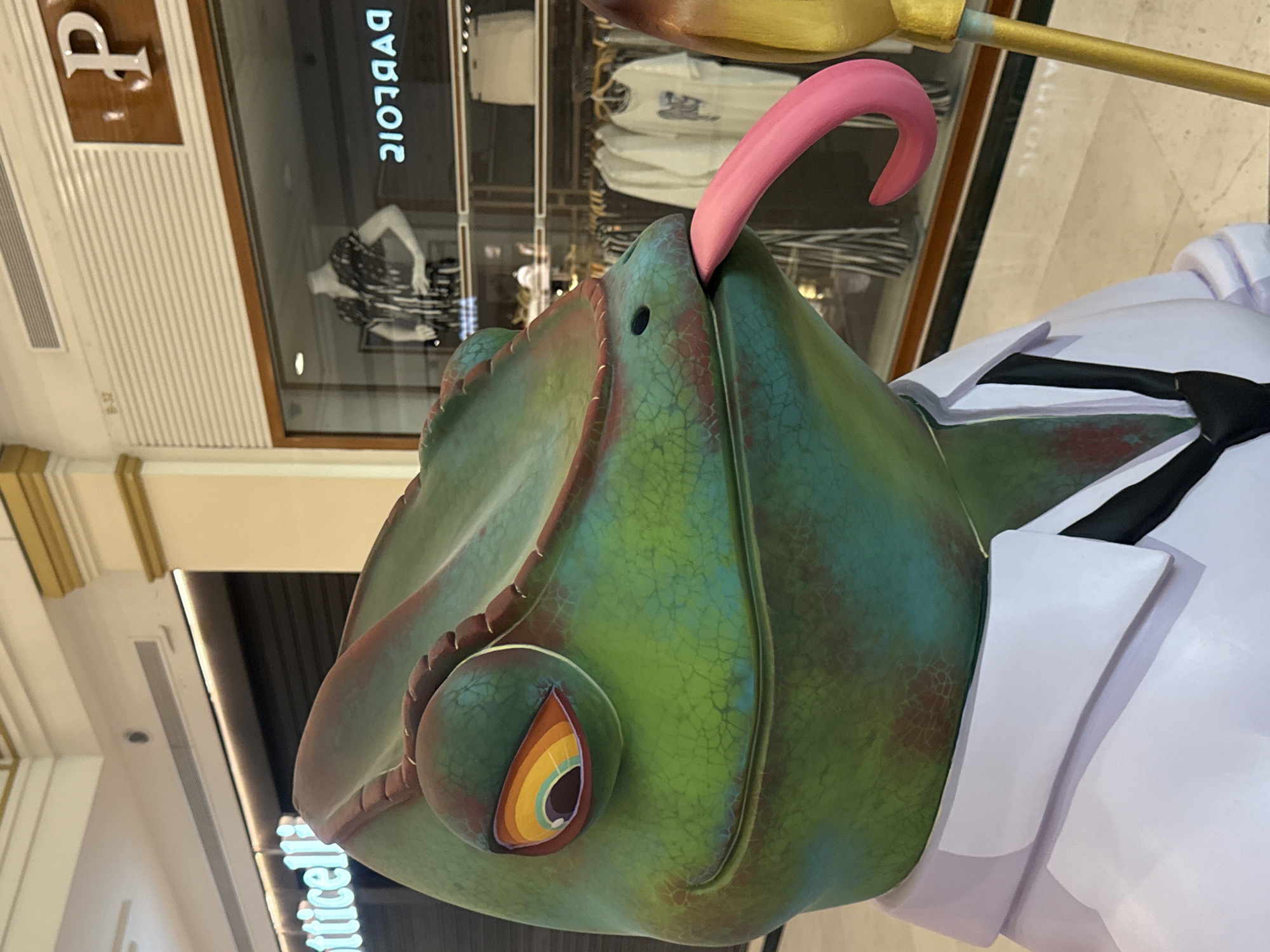 A life-sized, colorful statue of a chameleon wearing a white collared shirt and a black tie is displayed in what appears to be a shopping mall. The chameleon has a vibrant green and red mottled skin tone, a large eye with a rainbow-colored iris, and a long, pink tongue sticking out of its mouth. The background shows a storefront with mannequins and clothing racks visible through a glass window, as well as signage that includes "otticell" and "P" with "2107яад" reversed. The floor is tiled, and a golden pole with a yellow tip is partially visible to the right of the statue.