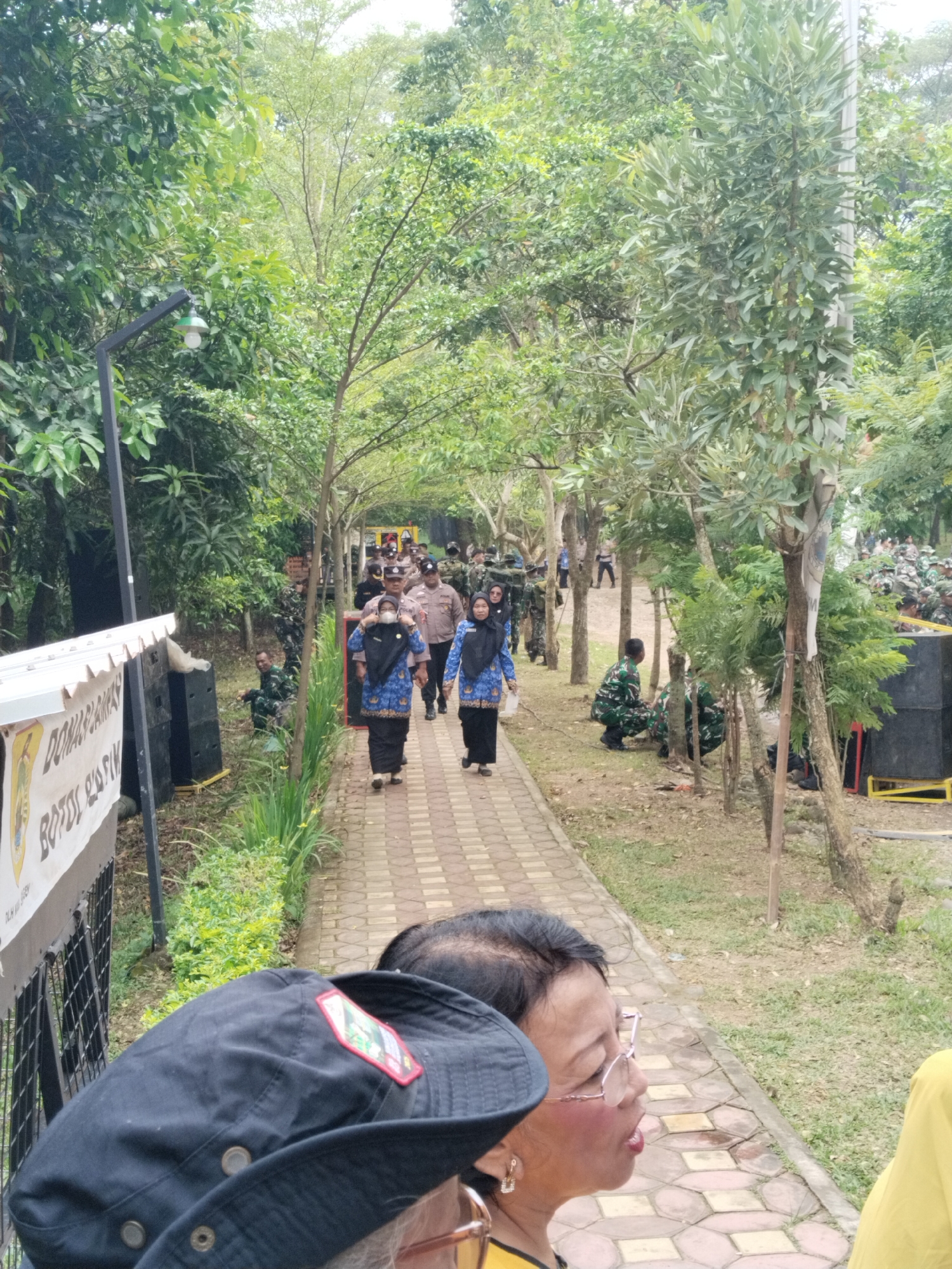 The scene captures a group of people walking along a brick pathway in what appears to be a park or outdoor area. A police officer in uniform is positioned in the center of the pathway, flanked by two women in blue patterned shirts and dark bottoms. Some individuals are wearing face masks. On either side of the pathway, there are trees, foliage, and black speakers. In the background, other individuals in military uniforms are present. In the foreground, there is a partial view of a person wearing a black hat with a patch. The overall atmosphere is one of an outdoor gathering or procession. The city and specific location are Unknown, Unknown.