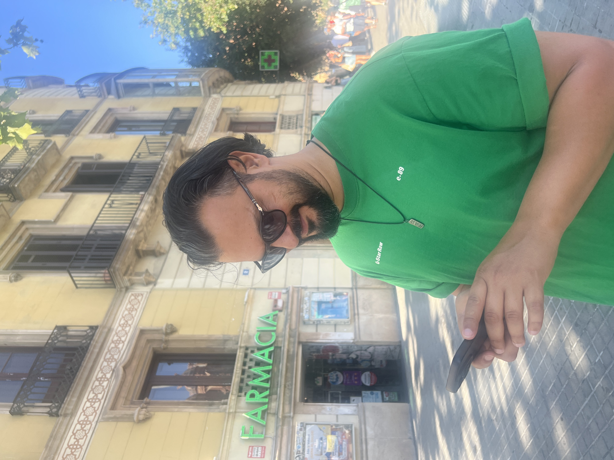 The image displays a man in a medium shot, situated on a city sidewalk in Barcelona, Spain, under bright daylight. The man, with dark hair, a beard, and wearing dark sunglasses, is looking down at a black smartphone held in both hands. He wears a bright green crew-neck t-shirt with "G-Star Raw" printed on the upper left chest and "e.89" on the lower right chest; his sleeves are rolled up. A black cord necklace with a metallic pendant, partially inscribed "RAW", is visible.
Behind the man, a multi-story building with a light yellow facade dominates the background. The building features ornate window frames, balconies with dark iron railings, and decorative friezes between floors. The ground floor houses a "FARMACIA" (pharmacy) identified by a prominent green neon sign and an illuminated green cross sign above its open entrance. A red alarm box marked "ALARMA 24H" and a small blue sign with the number "2" are affixed to the building's facade near the pharmacy.
To the right of the man, dense green foliage from trees partially obscures more of the building, which includes a bay window structure. Through the trees, another illuminated green cross pharmacy sign is visible in the distance, alongside a blurred crowd of people indicating a busy street or plaza. The sky above is clear blue. Strong shadows are cast on the light grey, patterned sidewalk, suggesting the sun is high and to the right of the frame.