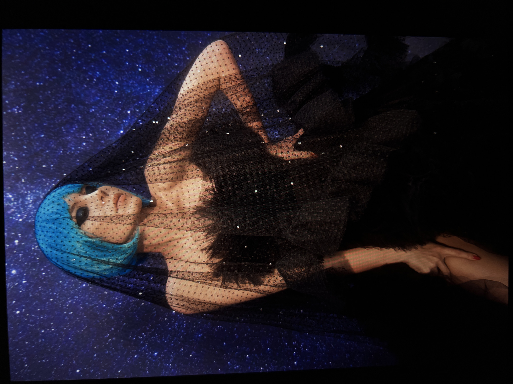 A person with bright blue hair and dark eye makeup is posing against a dark blue background with white speckles, resembling a starry night. They are wearing a sheer black veil with small polka dots and scattered sequins, and a black ruffled dress with a feathery texture. Their left arm is bent at the elbow, with their hand resting on their hip, while their right arm is extended downwards, with their hand resting on their leg. The lighting is dramatic, casting shadows on the person's face and body, and highlighting the sequins on their attire. The image does not depict any identifiable locations within the city of Saint Petersburg, Russia.