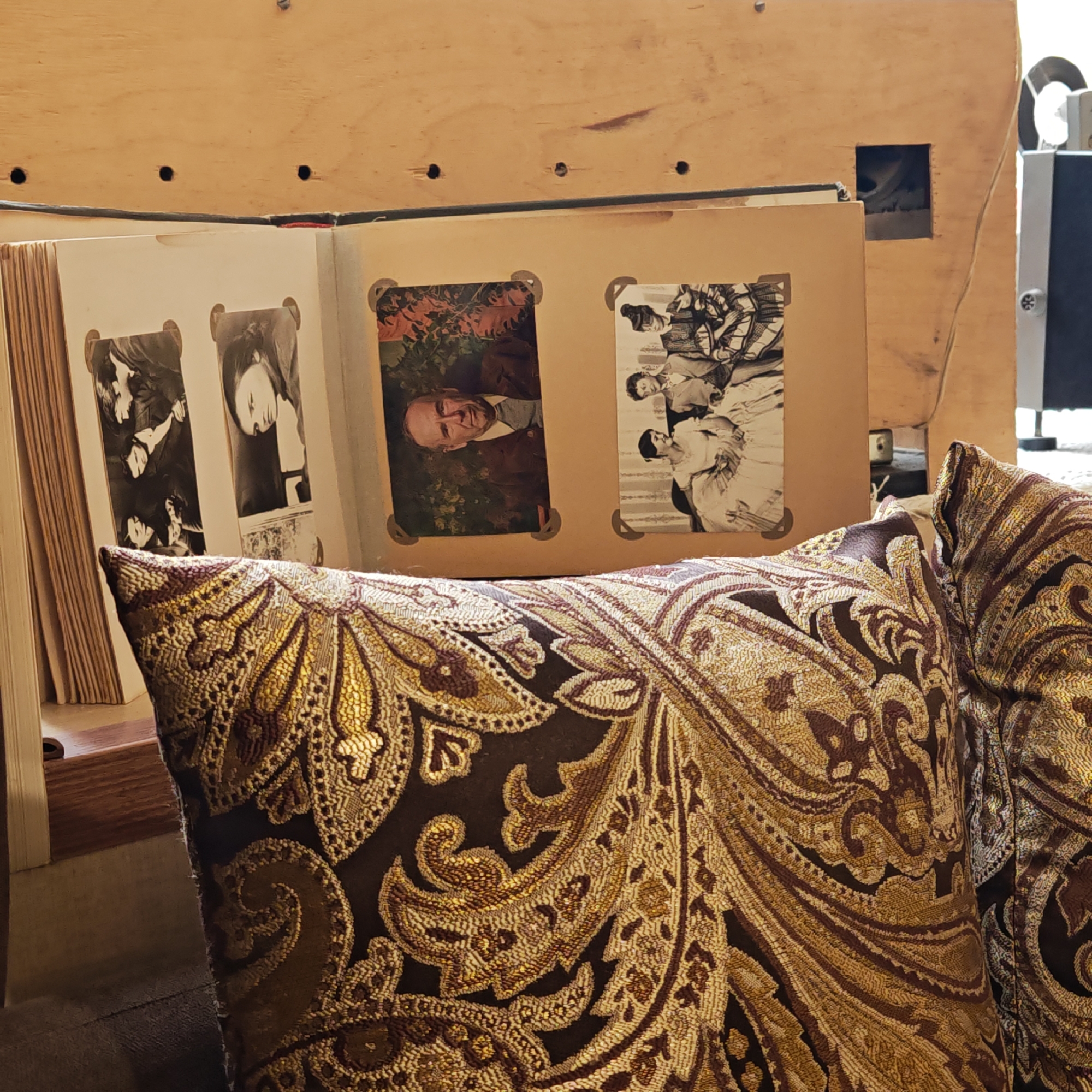 A close-up shot shows a partly opened photo album resting on patterned pillows. The album's pages display old black and white photographs, depicting people in various poses, including a man lying sideways. The pillows exhibit an intricate, ornate design in gold and brown hues. Behind the album, a light-colored wooden surface with drilled holes and a square cut-out can be seen, partially obscuring the background. The setting does not appear to be in Rostov, Russia.