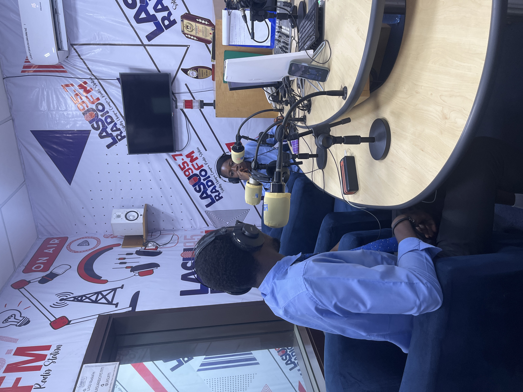 The image depicts a radio studio at LASU Radio 95.7 FM, a campus radio station situated in Ijanikin, Nigeria. The studio is equipped with a sound mixer, microphones, computer keyboard, and a television mounted on the wall. A banner featuring the radio station's logo, name, and the phrase "Solely Unique Campus Radio Station" is visible on the wall. Two individuals are present in the studio, seated behind the table, wearing headsets and positioned at microphones. The environment appears to be a professional broadcast setting.