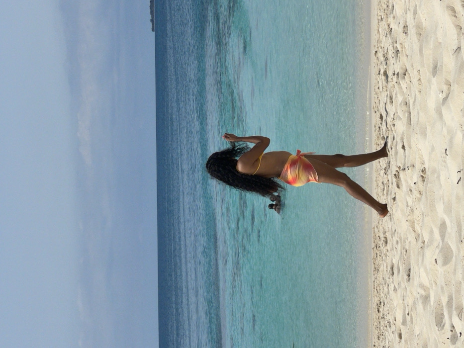 A pristine sandy beach environment is depicted in Zanzibar, Tanzania, under a bright sky. In the foreground, a person with long, dark, wavy hair walks barefoot from left to right. This individual is wearing a yellow bikini top and a translucent sarong with a red, orange, and yellow tie-dye pattern, tied at the waist. Their right hand is raised to the head, seemingly adjusting their hair. In the midground, the water is clear and turquoise, gradually deepening to a darker blue towards the horizon. Two indistinct human figures are partially submerged in the turquoise water on the left side of the midground. The far background shows the ocean meeting a light blue sky with minimal cloud cover, and a faint, hazy outline of a small island or landmass is visible on the far right horizon.