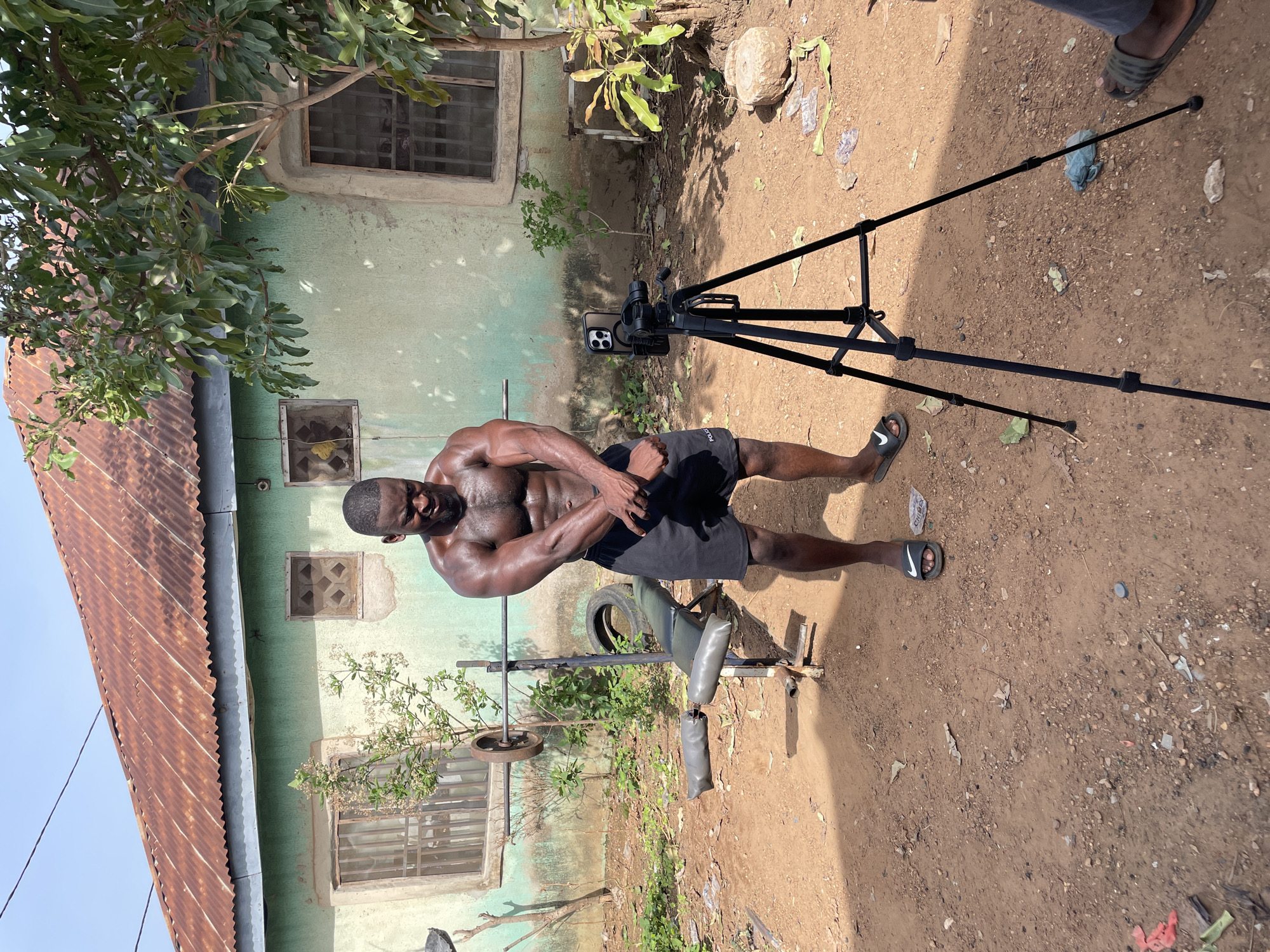 An African man with a muscular build is posing in a dirt backyard. He is dark-skinned, shirtless, wearing black shorts, and black and white sandals. He stands near a weathered weight bench and a tripod holding a smartphone. The backdrop features a partially rusted corrugated metal roof over a building with a green painted exterior and several barred windows. The yard contains sparse vegetation and debris. A second person's legs and feet are visible in the background near the tripod. The location is suspected to be in Akwanga, Nigeria.