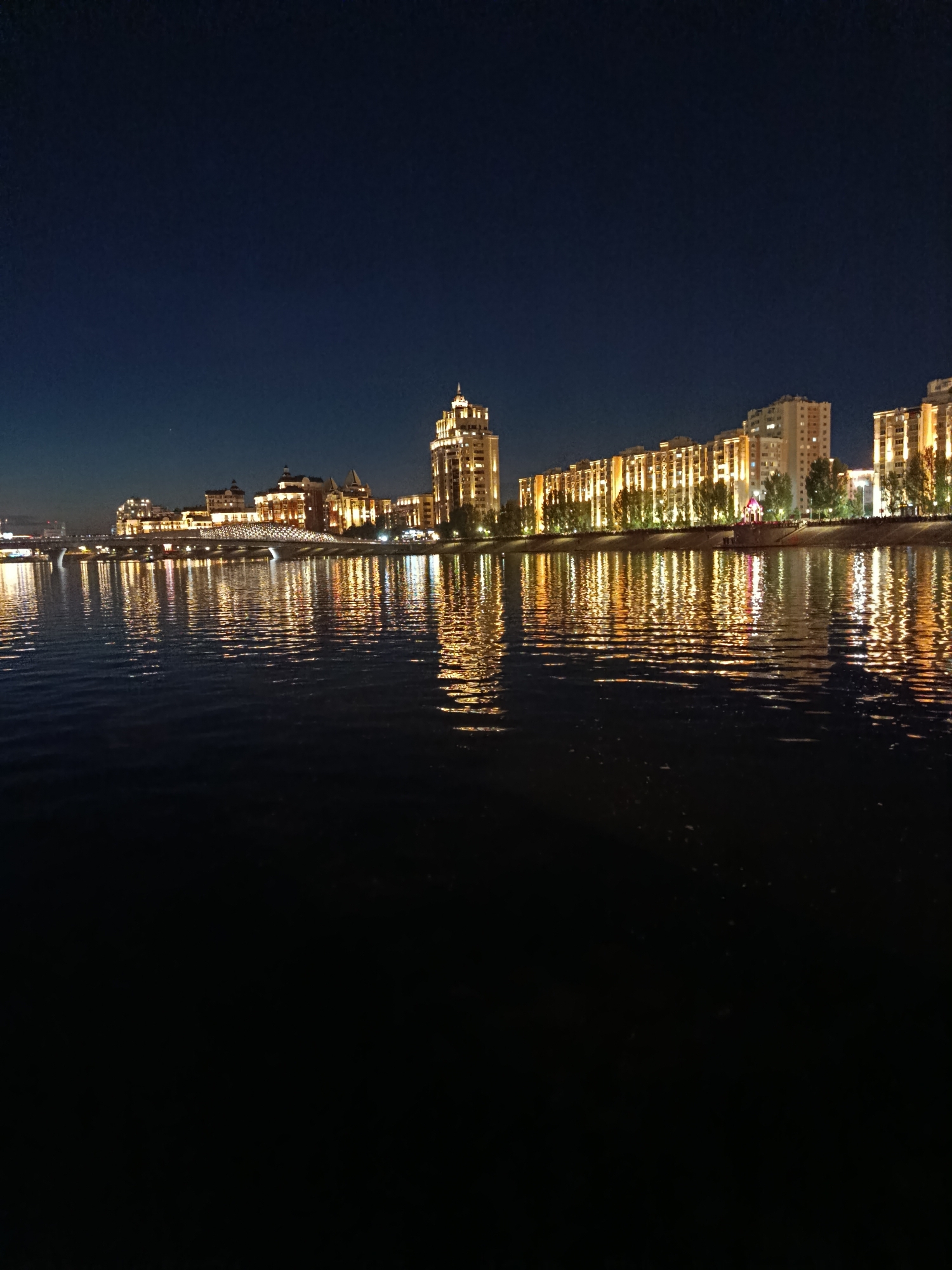 The image captures a night scene along the Ishim River in Astana, Kazakhstan. On the far bank, a line of modern buildings and apartment complexes are illuminated with warm, golden lights, creating a reflection on the dark water. A bridge with illuminated arches spans the river in the mid-ground, connecting the two sides of the city. The sky is a deep, dark blue, indicating the late hour. The river itself is calm, with ripples disturbing the reflections of the lights. The foreground of the image is almost entirely dark water, with the lights of the city dominating the background and mid-ground. There are no people visible in the image.