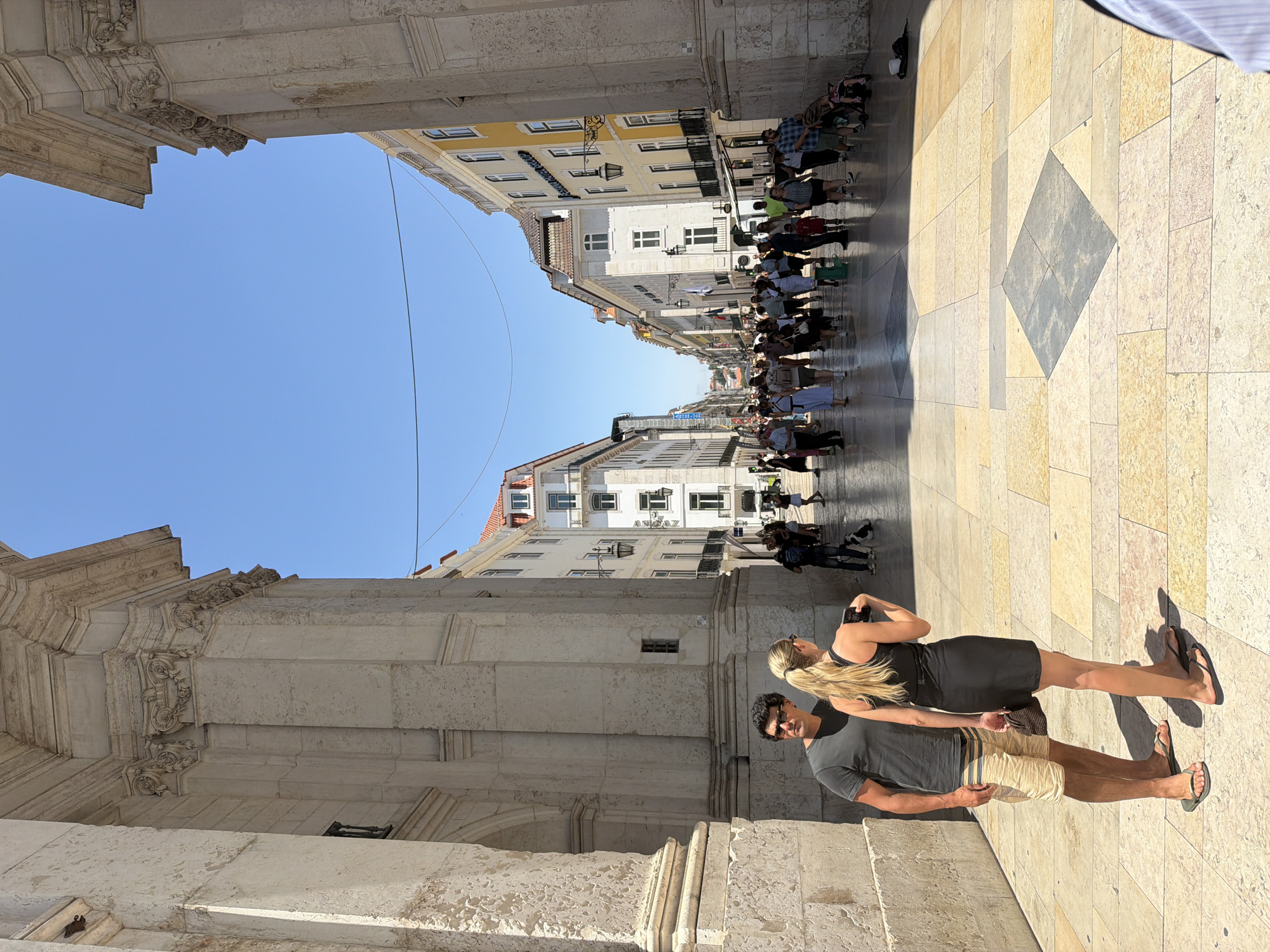 A sunny, wide, cobblestone street in Lisbon, Portugal, is shown from ground level. Two people, a man and a woman, stand in the foreground, facing away from the camera. The man wears a dark t-shirt and khaki shorts, and the woman wears a black dress. They are standing near a large, stone archway.
In the background, the street is lined with multi-story buildings with balconies and windows. A crowd of people walks along the street, some looking at shops, others conversing. The sky is clear and blue. The scene appears to be a popular tourist area, with many people enjoying the warm weather.