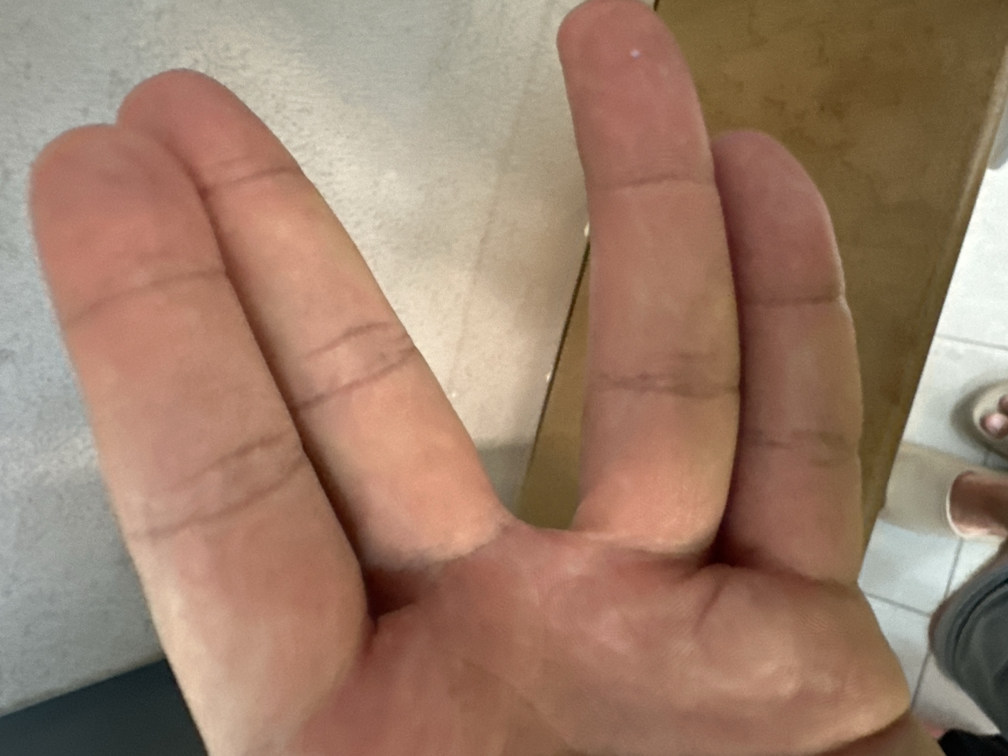 A close-up, overhead shot shows the palm and fingers of a person's hand, held over a textured, light-colored surface. The hand is positioned so that the fingers are fanned out slightly, with the index and middle fingers appearing to be held together. The skin tone is fair, and faint lines indicative of knuckles and finger joints are visible. The lighting casts soft shadows. The lower portion of the image reveals a person's legs, wearing gray shorts, and two light-colored slippers on their feet, suggesting they are indoors. The background behind the hand is a blurred, speckled surface, and a wooden countertop is visible below the hand and to the right. The scene appears to be in a residential setting within the city of Tivat, Montenegro, likely a home environment. There is a small white speck visible on the tip of the person's right index finger.