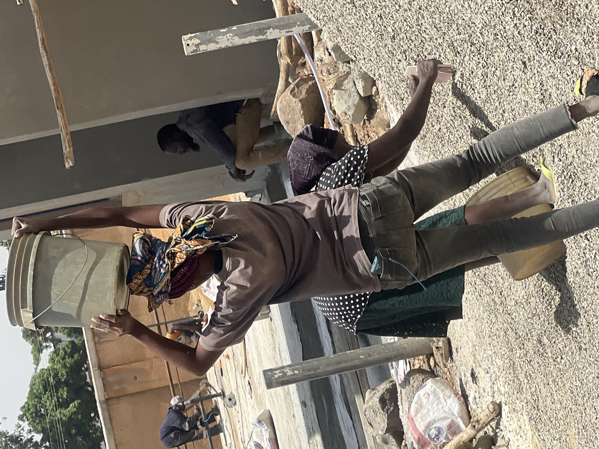 In Barakin Lerawi, Nigeria, two individuals are actively engaged in construction work on a building. One person is carrying a bucket while another is working on the building structure. The environment contains construction materials, including gravel, bags, and scaffolding.