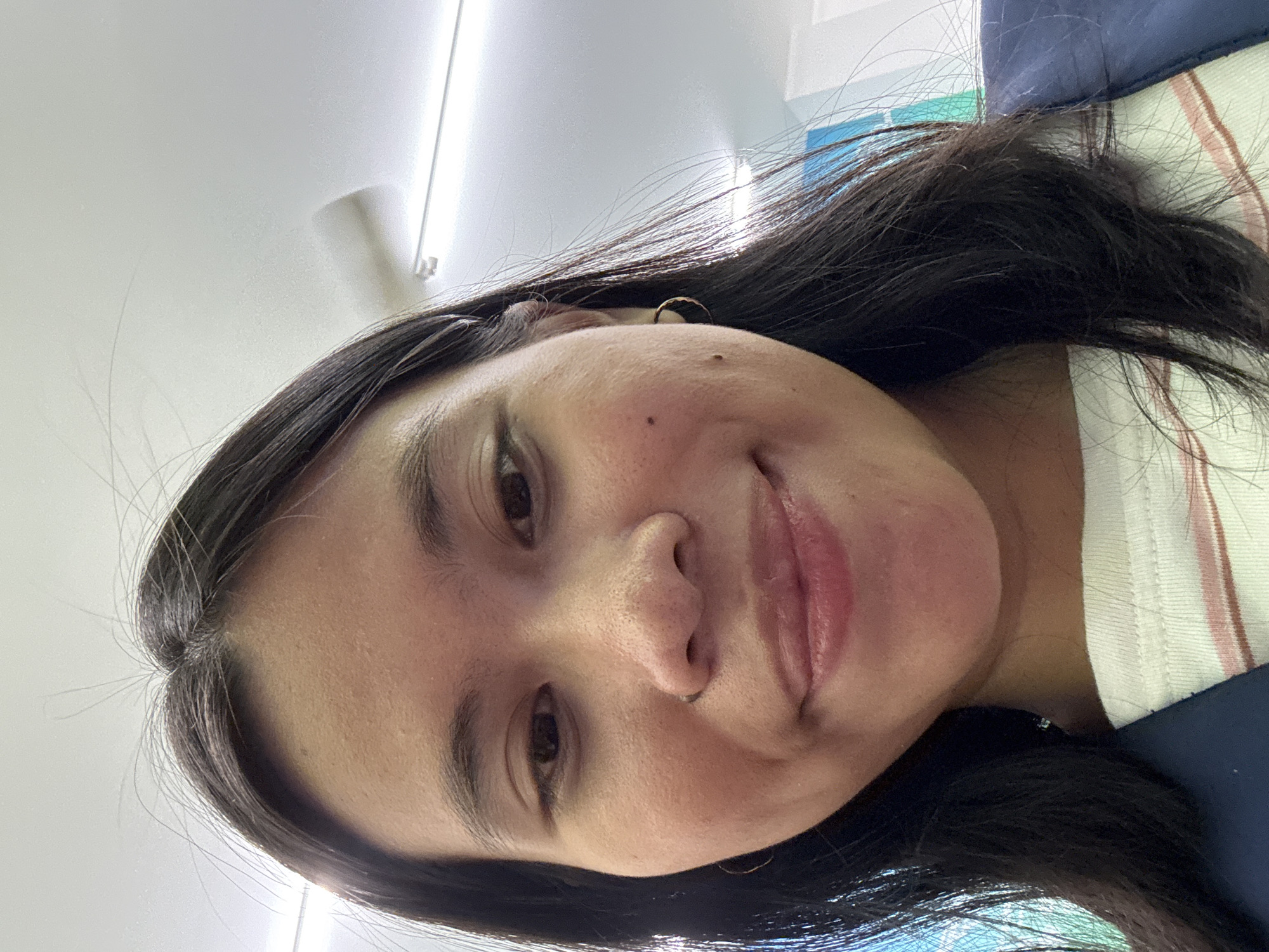 The image is a close-up selfie of a young woman, likely in an indoor setting. She has dark hair, dark eyes, and a light complexion. She is wearing a white t-shirt with thin horizontal stripes in pink and brown, and a dark blue garment, possibly a jacket or backpack, is visible on her shoulder. She is also wearing a small hoop earring in her right ear. The background shows a white ceiling with fluorescent lighting fixtures, suggesting an office, classroom, or similar public space. The lighting is bright, casting a slight glare on the ceiling. There are no specific landmarks or clues in the image that would definitively place it in East Jakarta, Indonesia.