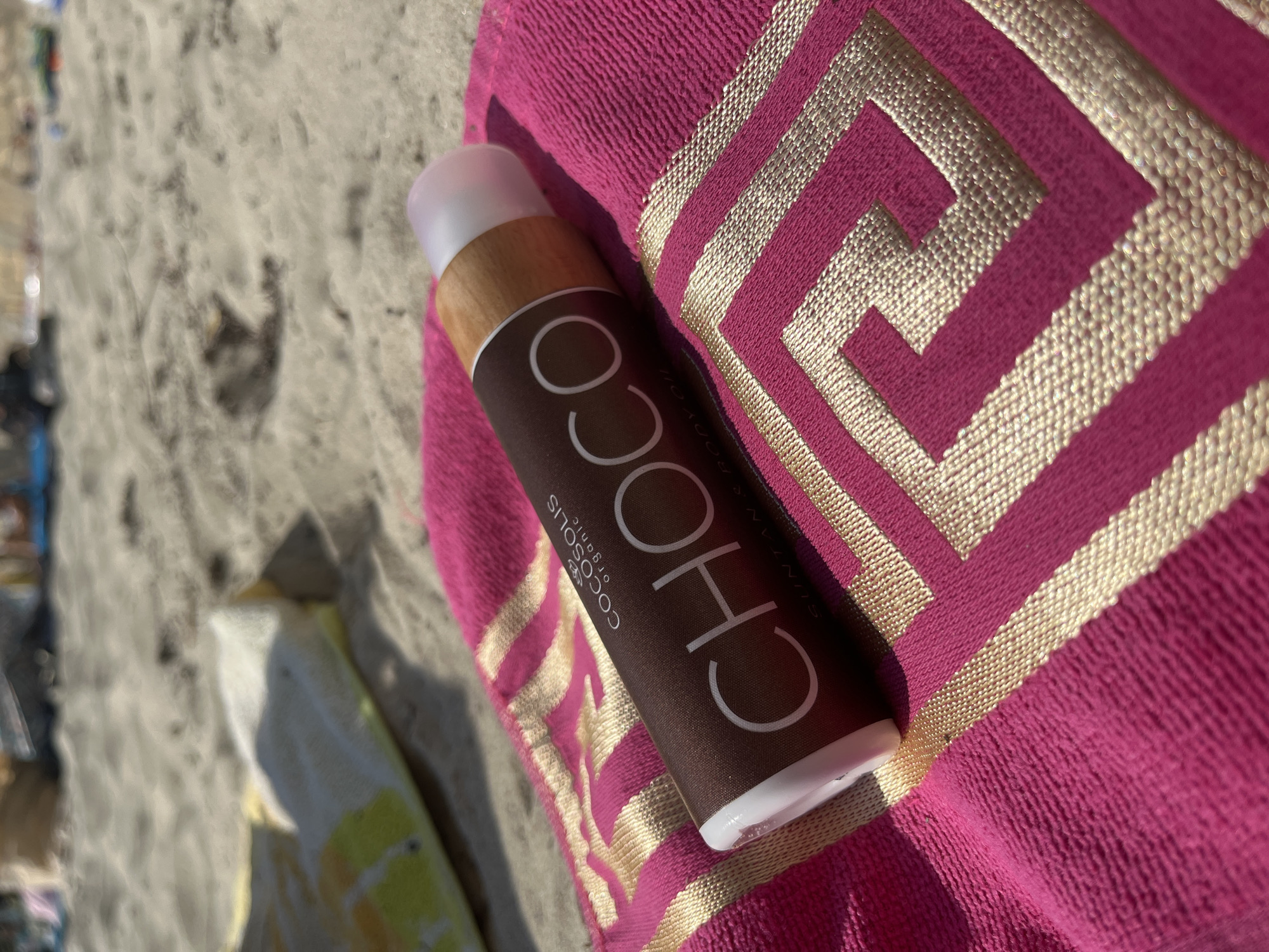 A close-up shot captures a product tube for "CHOCO" branded "COCOSOLIS organic" suntan and body oil. The tube is placed on a bright pink beach towel with a gold geometric pattern. The towel rests on a sandy beach, with some blurred beachgoers and a yellow and white object in the background. The scene suggests a sunny day at a beach, possibly in Nesebar, Bulgaria, given the context.