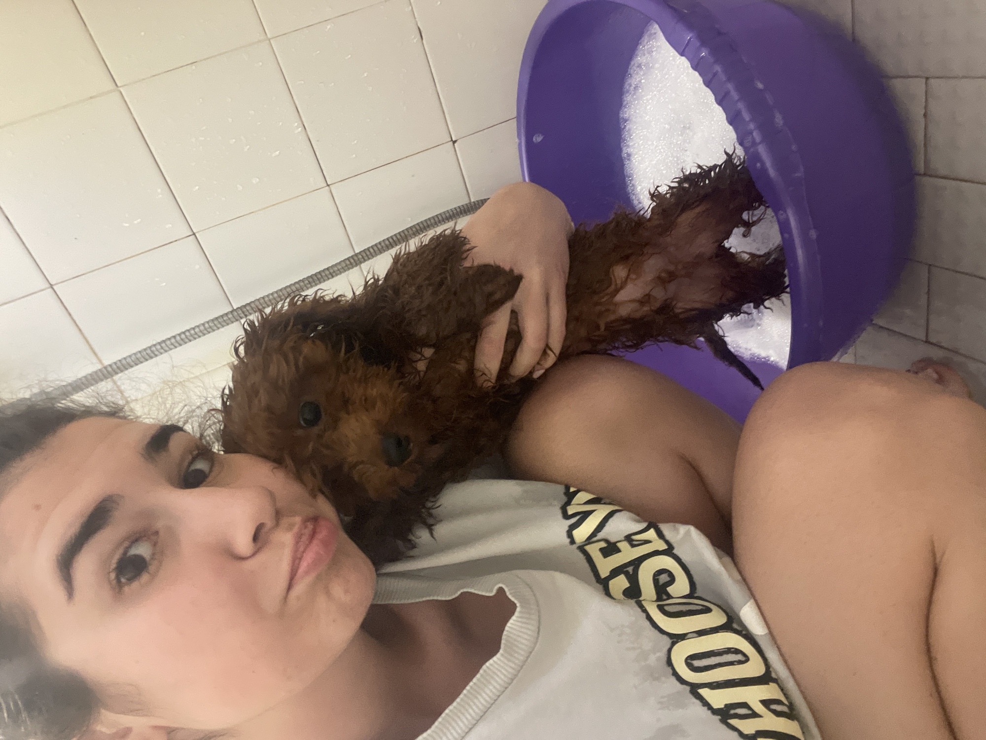 A woman takes a selfie with a wet brown dog in a bathroom with white tiled walls. She is holding the dog close to her chest. A purple plastic tub filled with soapy water is adjacent to the dog. The location cannot be determined from the given information and thus I cannot provide a description of Sofia, Bulgaria.
