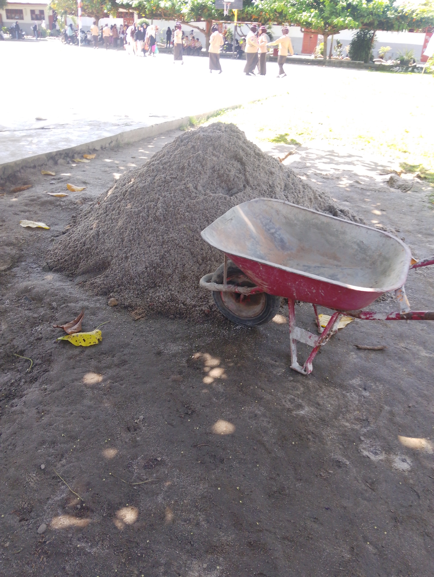 In the city of Serbelawan, Indonesia, a construction scene unfolds. A large pile of sand sits on the ground, likely for building purposes. Beside the sandpile, a red wheelbarrow is positioned, its metal basin filled with what appears to be more sand. The wheelbarrow's handles and frame are visible, along with its single wheel. The ground surrounding the sand and wheelbarrow is a mix of dirt and sand. In the background, a group of people, some wearing light-colored uniforms, are gathered. The setting appears to be an outdoor area, possibly a schoolyard or a public space, with buildings and trees visible in the distance. The sun is shining, casting shadows on the ground.
