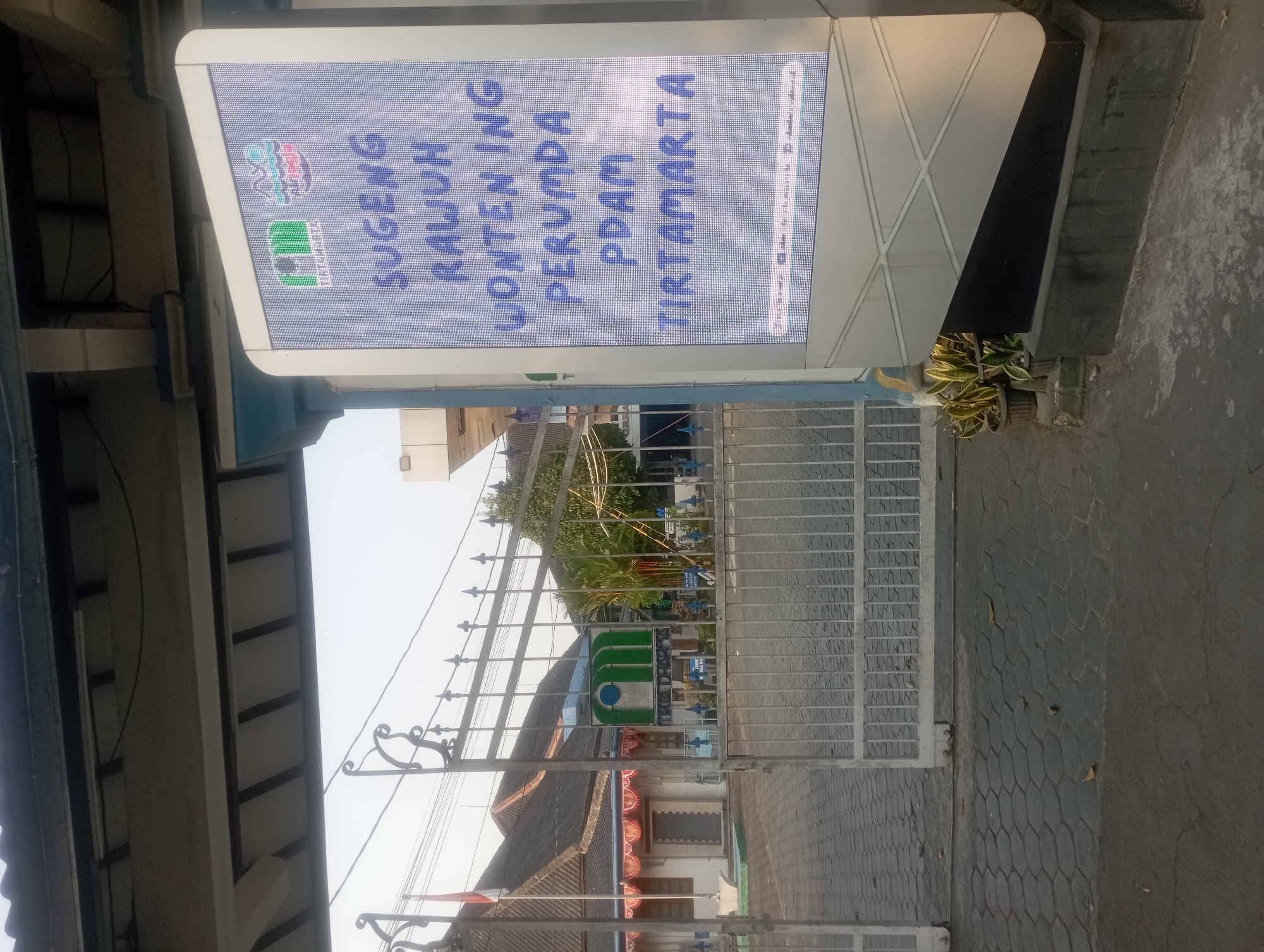 The image shows an outdoor scene with a large, illuminated digital sign on the right and a building with a gate in the background on the left. The digital sign displays text in a Javanese script, including "Sugeng Rawuh Wonten Ing Perumda" and "Tirtamarta," which translates to "Welcome to Perumda Tirtamarta." Perumda Tirtamarta is the regional water utility company of the Special Region of Yogyakarta, Indonesia. The sign is positioned at the entrance to a facility, with a paved walkway and a metal gate in front of a building. The building appears to be an office or public institution, with traditional Javanese architectural elements such as a tiled roof and ornate details. The overall environment suggests a public space in Yogyakarta, Indonesia, likely the entrance to a Perumda Tirtamarta office. There are no people visible in the image, and the focus is on the signage and the entrance to the facility.