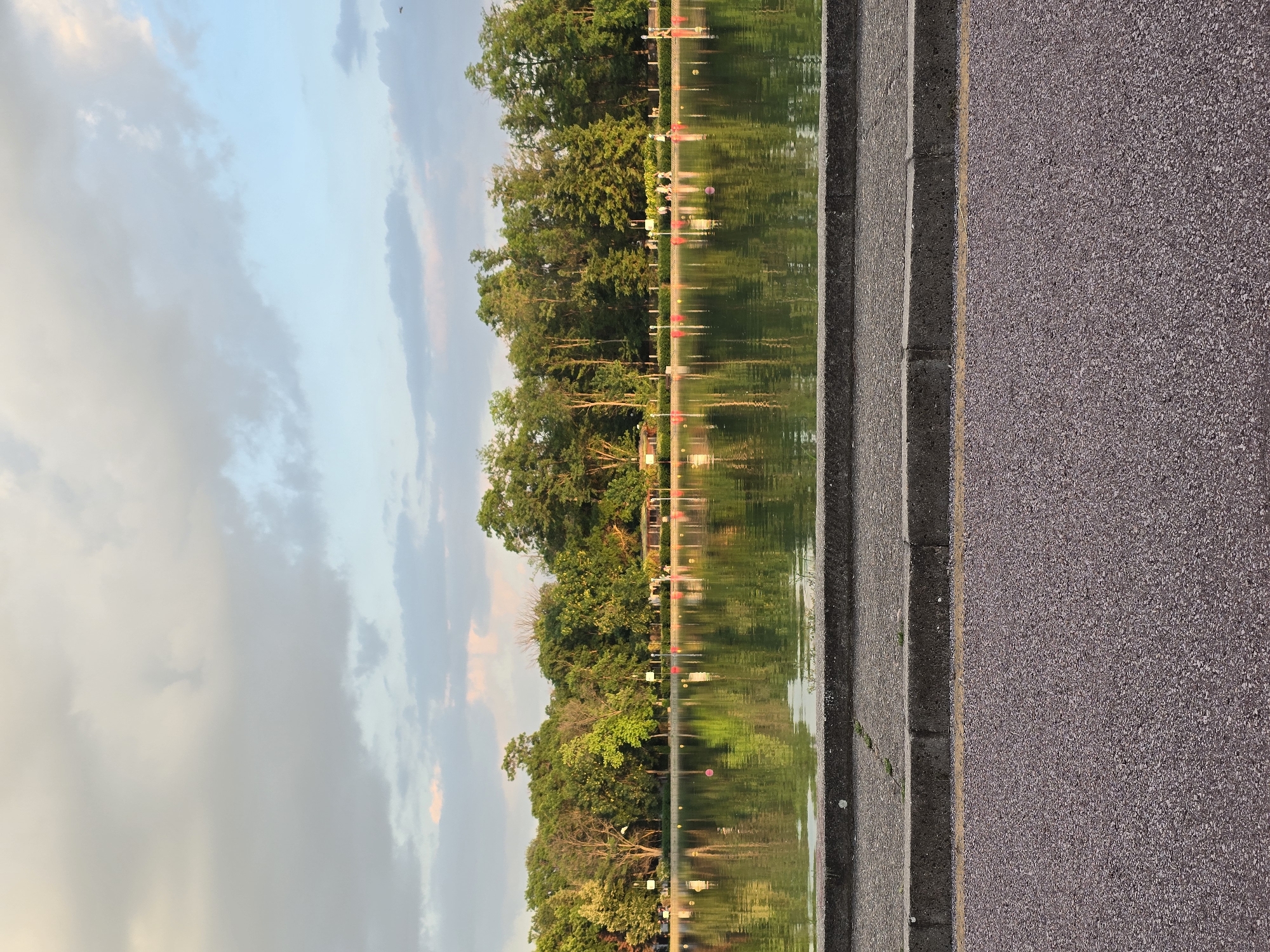 The image shows a body of water reflecting trees and sky. The right edge of the image features a paved surface, possibly a road or walkway, with a concrete curb. The sky exhibits variations in cloud cover and coloration, suggesting either early morning or late evening light. The water's surface is calm, facilitating clear reflections. The location appears to be a park or recreational area, indicated by the presence of trees and water. The lack of people or buildings indicates a natural or landscaped environment. The city is not identifiable from the visual data.