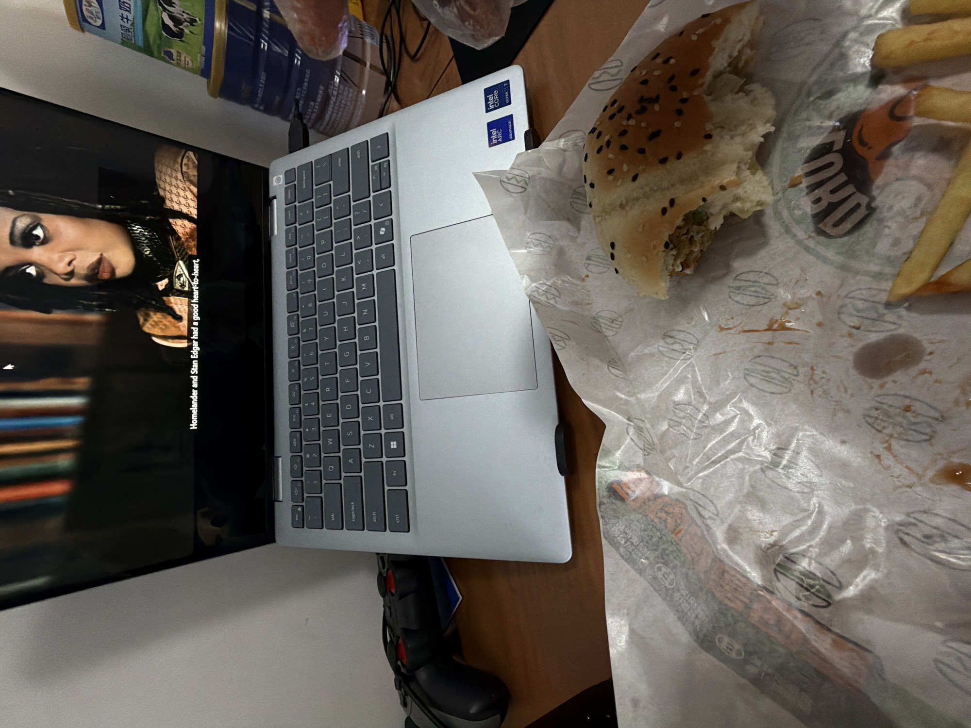 The image displays an indoor scene in Jiangning District, China, focusing on a casual meal and entertainment setup. A silver laptop, open on a brown desk or table, serves as a central element. The laptop features "Intel Core Ultra 7" and "Intel Arc Graphics" stickers prominently displayed on its palm rest. Its screen is either displaying content or connected to a larger television screen positioned vertically to the left of the laptop.
The television screen shows a close-up of a woman with dark hair and striking eye makeup, presumably from a show or movie. Subtitles are visible at the bottom of the screen, reading: "Homelander and Stan Edgar had a good heart-to-heart," which is a line from the TV series *The Boys*.
To the right of the laptop, a partially eaten burger with sesame and black sesame seeds on its bun, along with several french fries, rests on grease-stained white wrapping paper. The paper features a recurring logo of a stylized burger, indicating fast-food packaging. A dark logo with text, possibly "BURU," is also visible through the wrapping paper near the fries.
In the upper background, stacked blue and white containers are visible, one of which clearly shows the Chinese characters "乳牛奶" (Rǔ niúnǎi), meaning "Milk," confirming a local context. A black and red gaming controller is partially visible beneath the laptop, suggesting additional leisure activities. The overall scene depicts a relaxed setting, combining eating a fast-food meal with watching media.